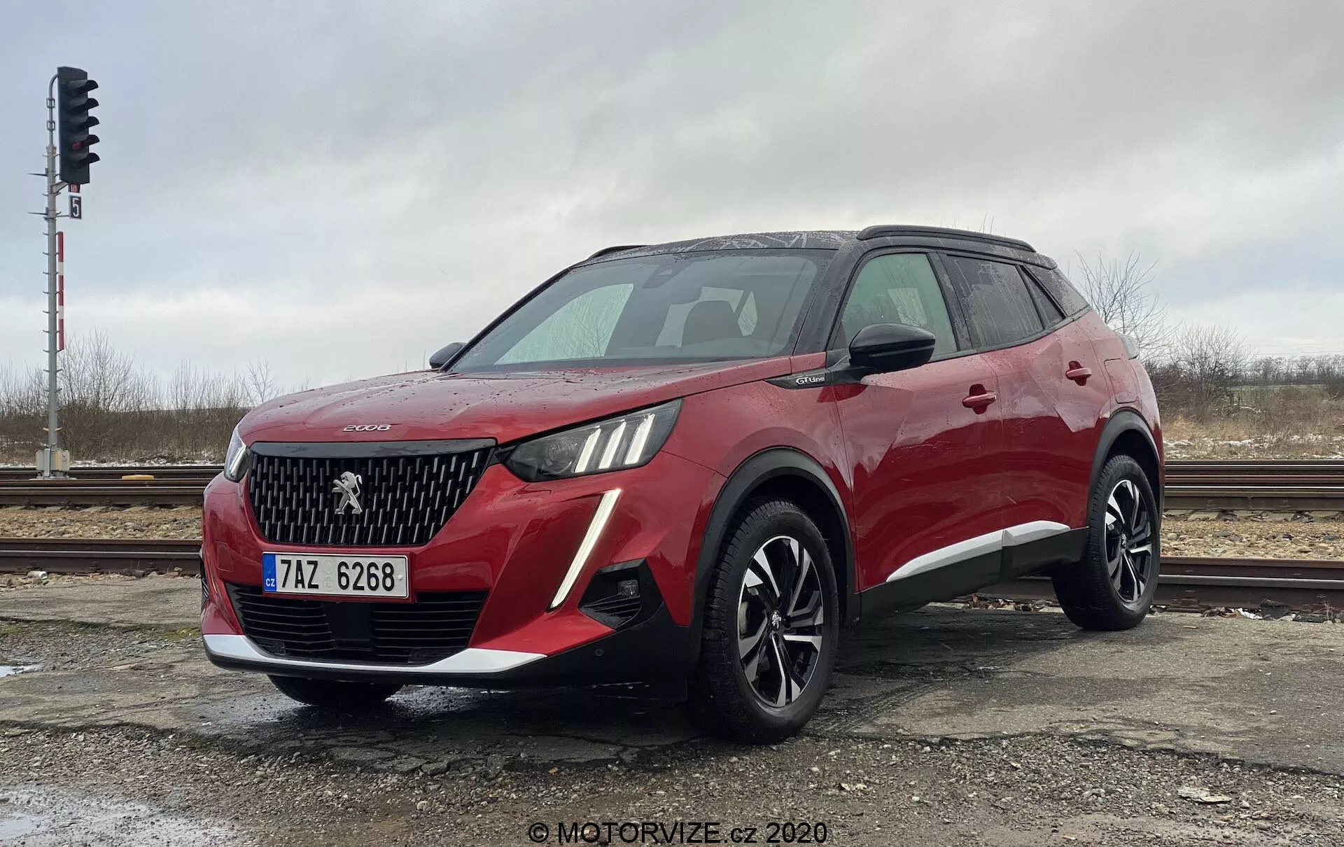 Trzy czwarte przedniego widoku nowoczesnego, czerwonego crossovera SUV Peugeot 2008 1.2 PureTech 100 Active. Ujęcie pod niskim kątem podkreśla wysokość i postawę pojazdu. Przód pokazuje wyraźny grill z siatką w kształcie rombu, emblemat prawdopodobnie reprezentujący lakiernik, oraz charakterystyczne reflektory z wbudowanymi światłami do jazdy dziennej LED. Przedni zderzak zawiera światła przeciwmgielne lub wloty powietrza. Profil boczny ujawnia aerodynamiczne linie, duże wieloramienne felgi aluminiowe, relingi dachowe oraz lusterka boczne z wbudowanymi kierunkowskazami. Zdjęcie uchwyciło design i technologiczne cechy tego SUV-a w pochmurny dzień, zaparkowany koło torów kolejowych.