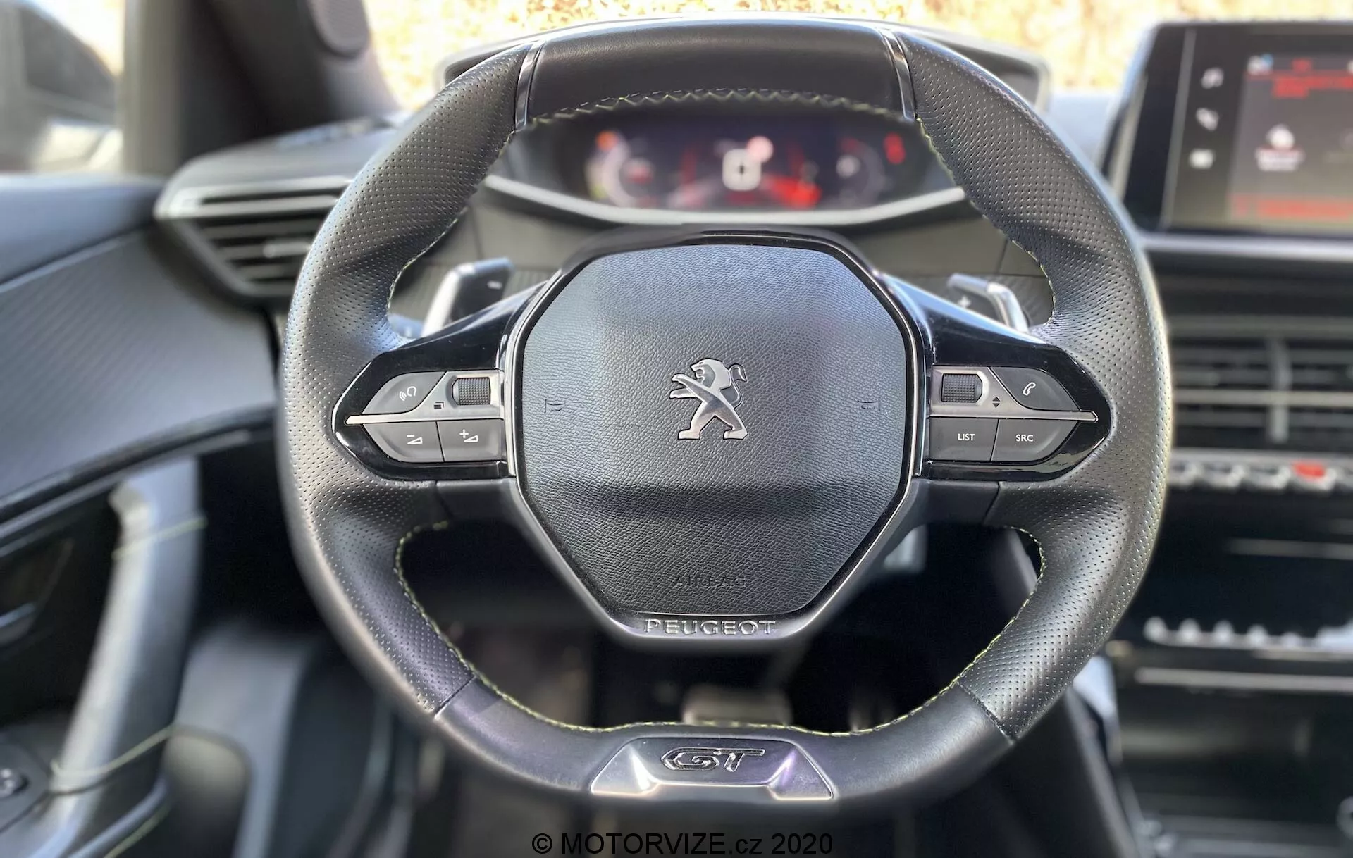 Wnętrze Peugeot 2008 2020 z widokiem na kierownicę i deskę rozdzielczą z perspektywy kierowcy. Główne cechy obejmują skórzaną kierownicę trzyramienną z wielofunkcyjnymi przyciskami, emblemat lwa Peugeot oraz konfigurowalny cyfrowy panel instrumentów. Widoczne elementy to ekran dotykowy systemu infotainment, kratki wentylacji i sterowniki konsoli centralnej z miękkim dziennym oświetleniem podkreślającym szczegóły kierownicy.