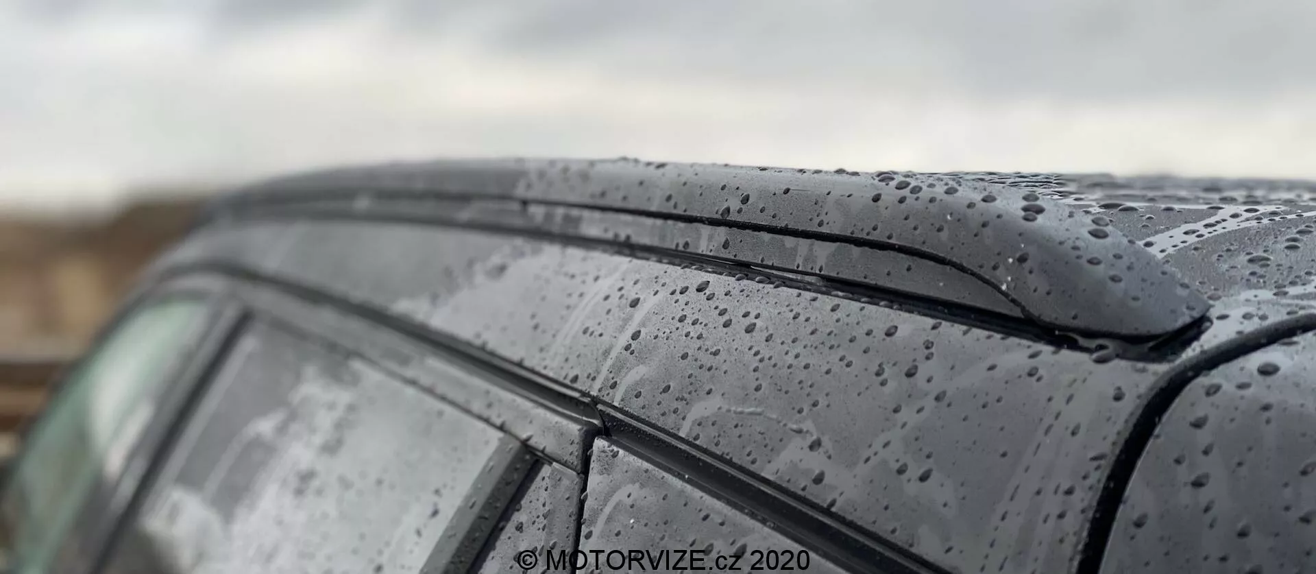 Zbliżenie na dach i boczne okno modelu Peugeot 2008 1.2 PureTech 100 Active 2020, podkreślające krople deszczu na eleganckiej powierzchni. Zdjęcie ukazuje ciemną, błyszczącą lakierowaną powierzchnię z kroplami deszczu oraz minimalistyczny design górnej krawędzi bocznego okna. Płytka głębia ostrości akcentuje teksturę zewnętrzną samochodu, rozmywając tło i części dachu, prezentując artystyczne ujęcie samochodu w mokrych warunkach.