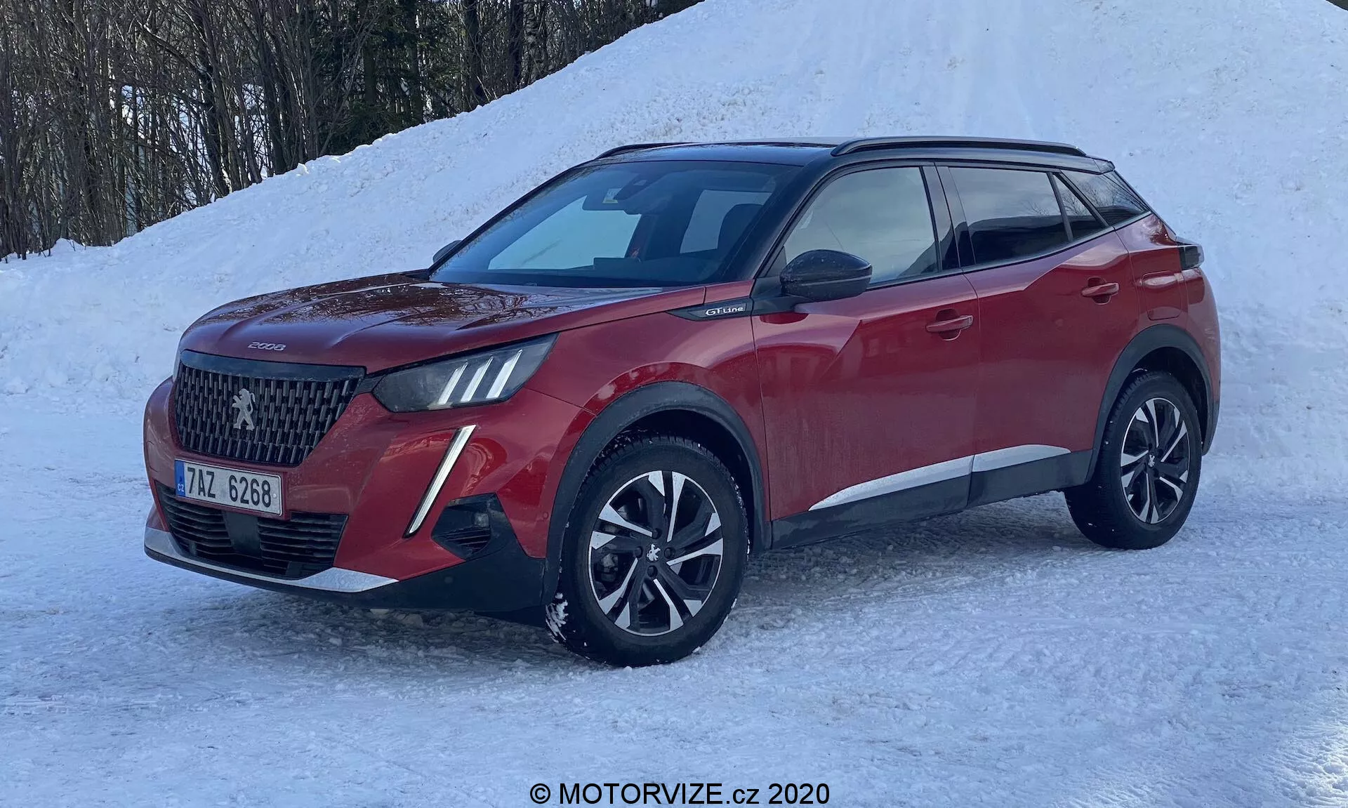 Nowoczesny SUV Peugeot 2008 1.2 PureTech 100 Active 2020 z trzech czwartych frontu, podkreślający dużą kratkę w romby, reflektory LED z DRL, wieloramienne felgi aluminiowe i rzeźbione czerwone metaliczne nadwozie. Ustawiony na zaśnieżonym podłożu, sugerujący zimowe możliwości prowadzenia, takie jak napęd na wszystkie koła.