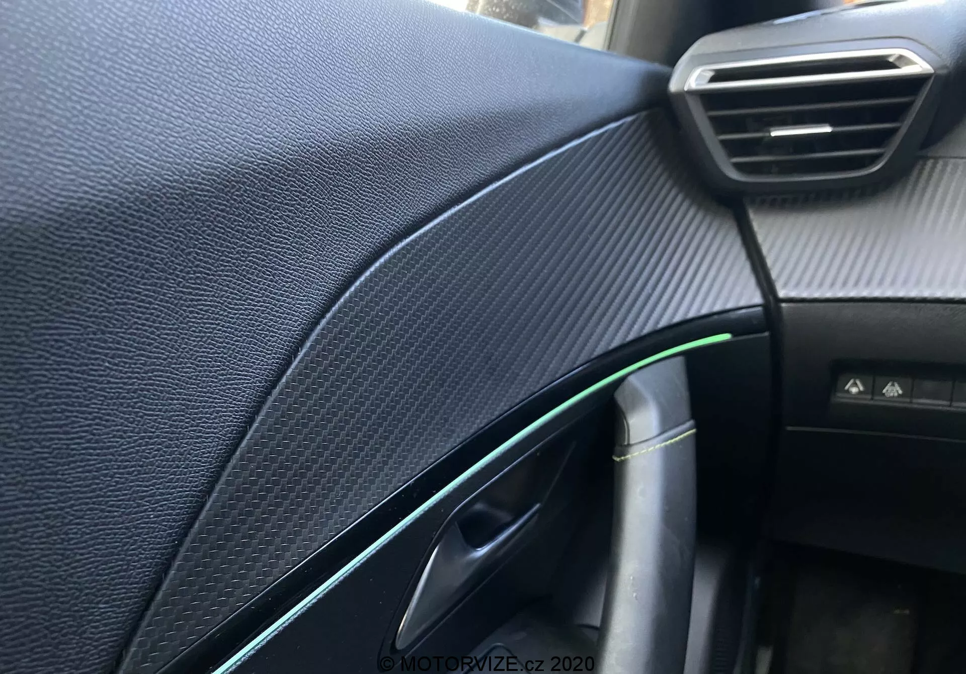 Zbliżenie wnętrza deski rozdzielczej i szczegółów od strony pasażera w Peugeot 2008 2020, koncentrując się na teksturowanej czarnej syntetycznej desce przypominającej włókno węglowe, widocznej regulowanej wentylacji, częściowo otwartych drzwiach z zielonym odbiciem i chromowanym akcentem, klamce drzwi, kablu z wtyczką ładującą, oświetleniu kabiny i niezidentyfikowanym przycisku, uchwyconych przy płytkiej głębi ostrości i naturalnym oświetleniu.