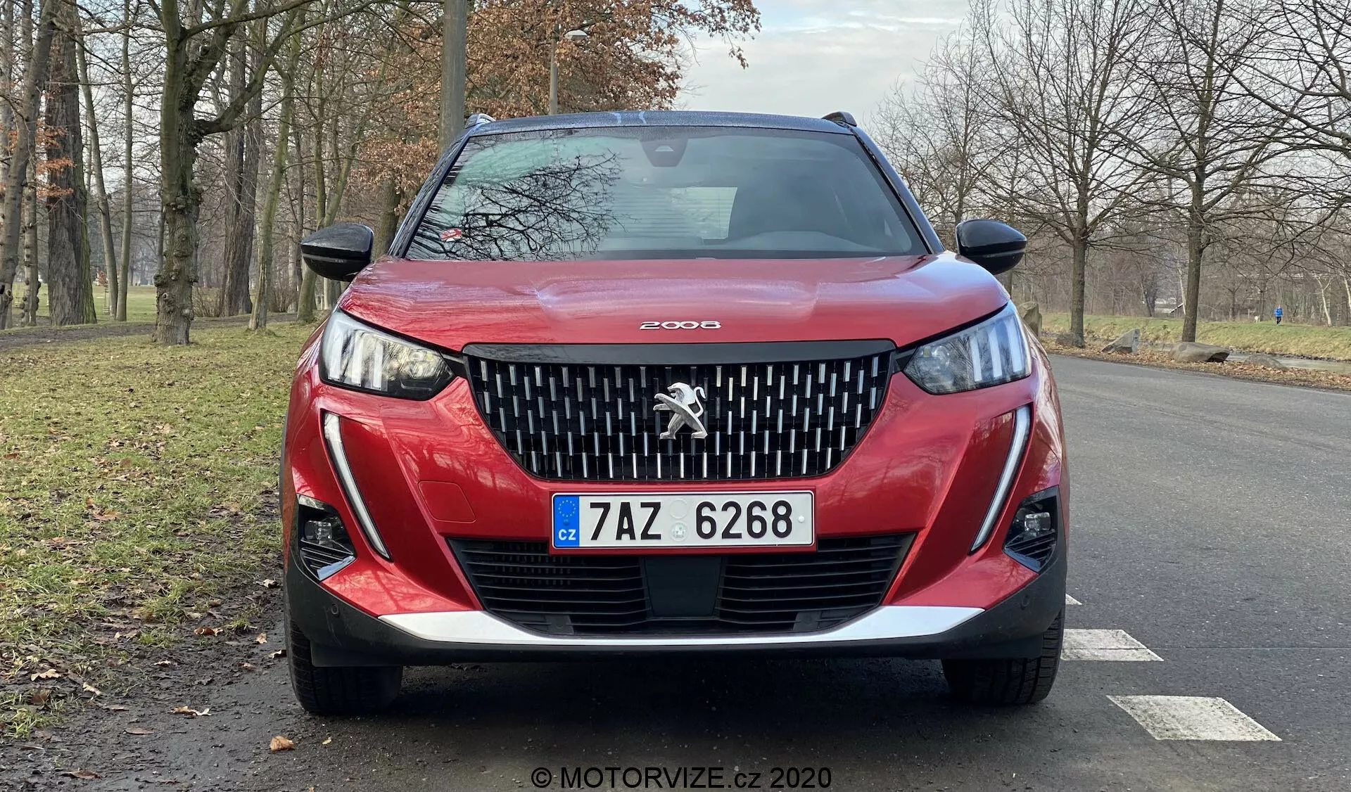 Zbliżony widok z przodu czerwonego Peugeota 2008 z rocznika 2020 (Peugeot-2008-1-2-PureTech-100-Active), ukazujący szczegółowy grill z wyeksponowanym lwem, agresywnie zaprojektowane reflektory z dzienne światła LED w kształcie 'kłów', rzeźbiony dolny zderzak i czeską tablicę rejestracyjną. Kontur maski dodaje dynamiki, a zdjęcie ukazuje częściowy widok aluminiowych obręczy i bocznych lusterek na suchej asfaltowej drodze z bezlistnymi drzewami i trawiastym terenem w tle, sugerując spokojne, naturalne otoczenie, być może w okresie jesiennym lub zimowym.