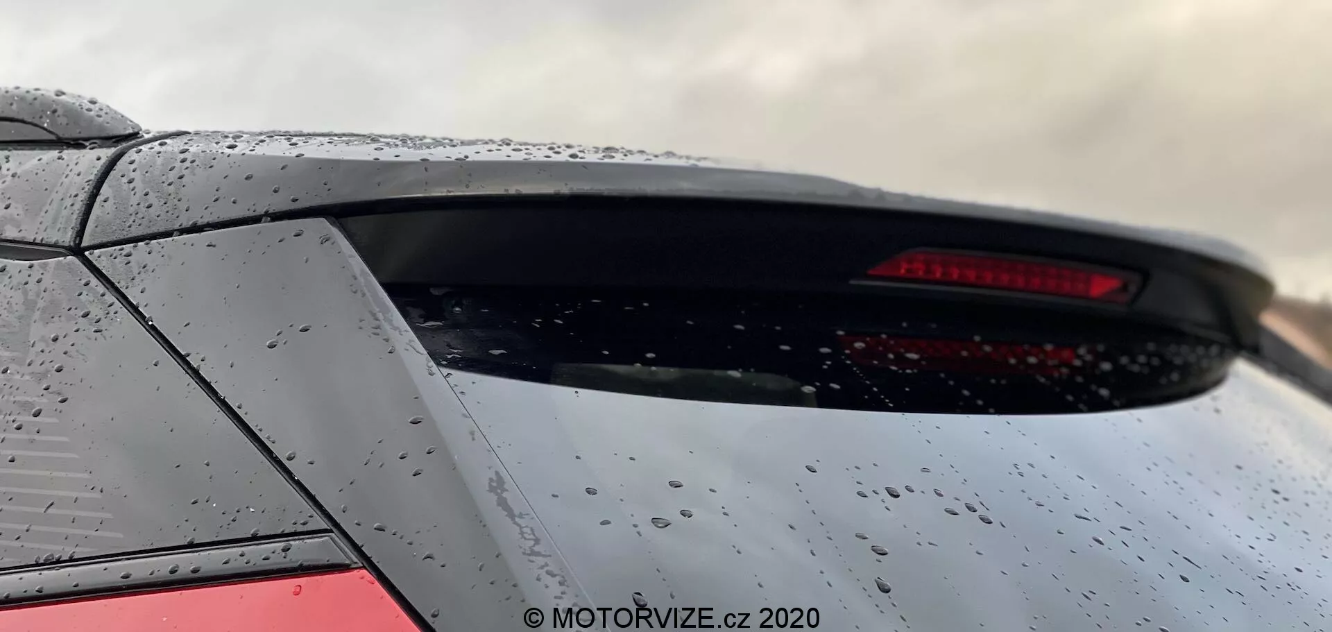Zbliżenie tylnej części modelu Peugeot 2008 z 2020 roku, podkreślające dolną część tylnej szyby i górną część klapy bagażnika. Kąt z dołu do góry podkreśla tylne okno hatchbacka, wbudowany tylny spoiler i tylne światło LED. Metalicznie szary samochód ma krople wody, co sugeruje niedawny deszcz. Obraz przycięty nie zawiera identyfikacyjnych logo.