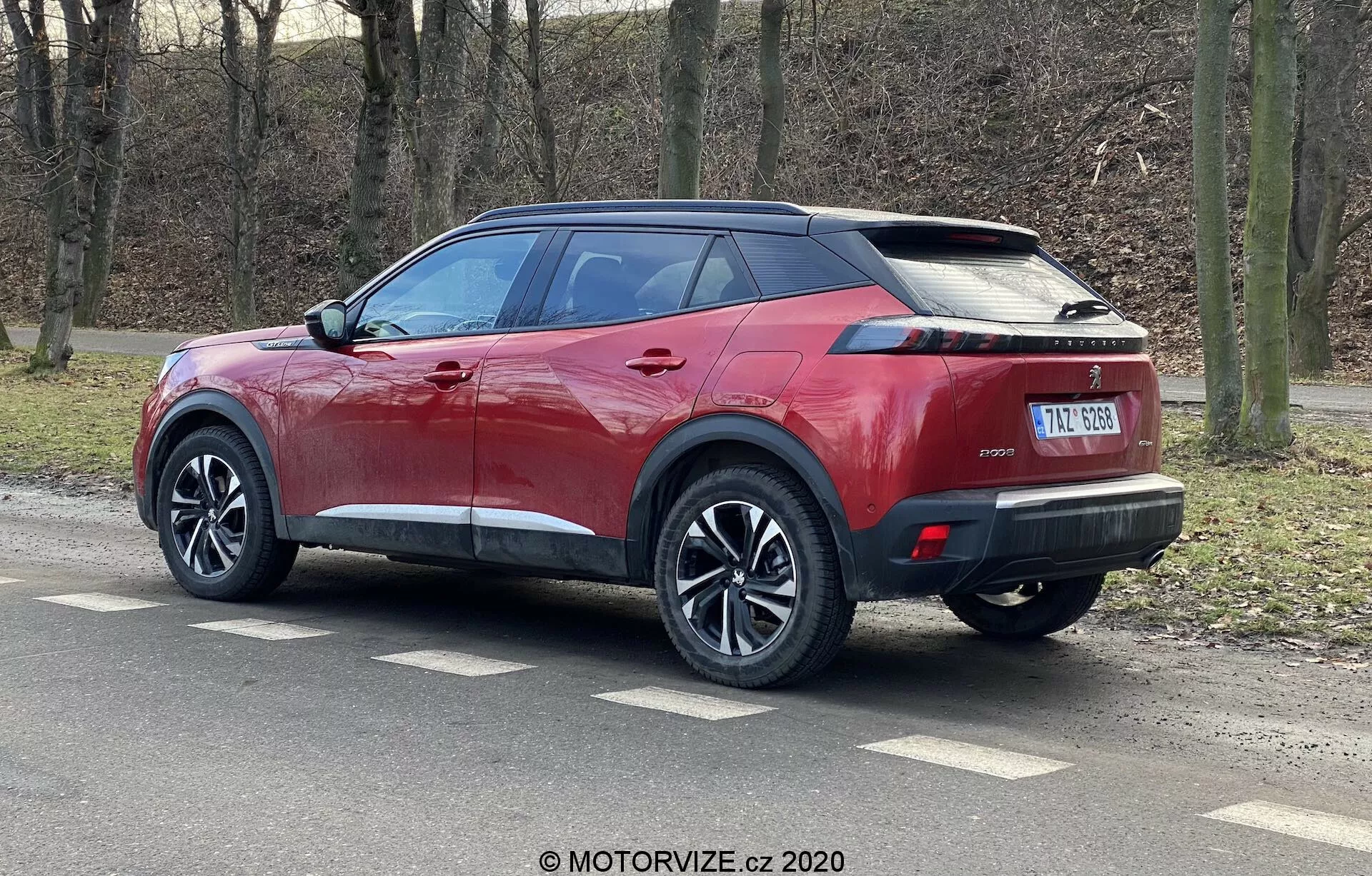 Obraz przedstawia nowoczesny SUV Peugeot 2008 2020 w jasnym metalicznym czerwonym kolorze z kontrastującym czarnym dachem, wieloszprychowymi kołami i oponami o niskim profilu. Zdjęcie wykonane z widoku trójkątnego tylnego, pokazuje ostre linie boczne, zintegrowany spojler, antenę w kształcie płetwy rekina, wąskie poziome tylne światła LED oraz elegancki tylny zderzak z czujnikami parkowania. Pojazd ma klamki i lusterka boczne w kolorze nadwozia z zintegrowanymi kierunkowskazami, przyciemniane tylne szyby oraz centralną tablicę rejestracyjną poniżej potencjalnego miejsca na tylną kamerę.