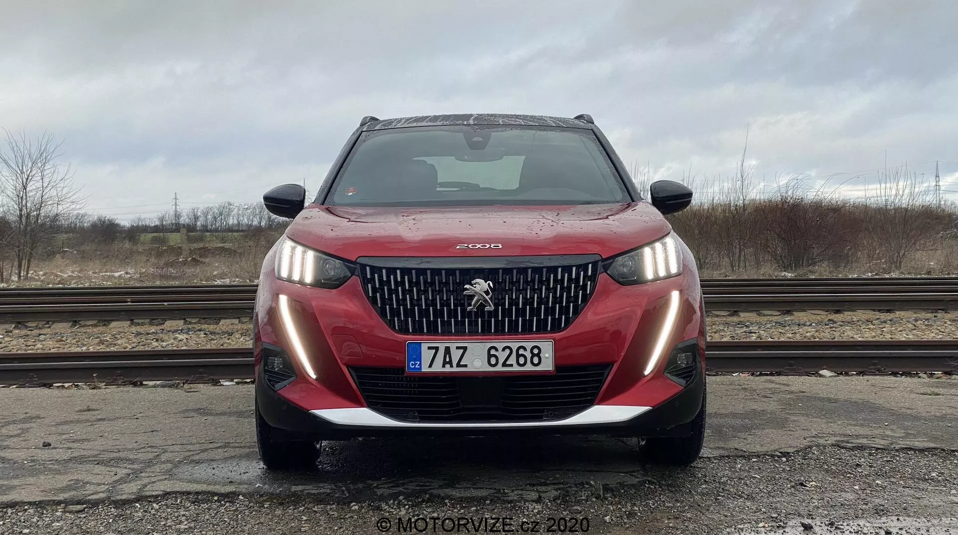 Widok frontalny czerwonego samochodu Peugeot 2008 2020 PureTech 100 Active, sfotografowanego z niskiego kąta, co podkreśla przednią kratkę, zderzak, reflektory i emblemat Peugeot. Samochód jest czysty, z kroplami wody na masce, przezroczysta szyba przednia odbijająca pochmurne niebo. Światła postojowe są włączone, widoczna jest europejska tablica rejestracyjna. Otoczenie obejmuje mokry grunt, tory kolejowe, nagie drzewa i szare niebo, co tworzy przemysłową atmosferę.