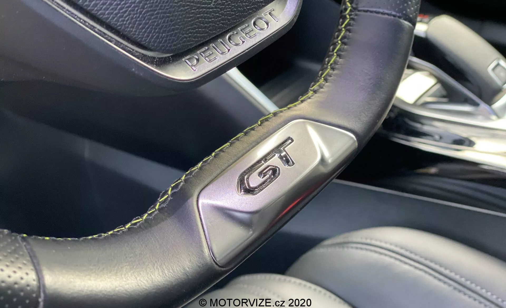 Zbliżenie wnętrza Peugeot 2008 2020, podkreślające znaczek 'GT' na perforowanej skórzanej kierownicy z zielonymi przeszyciami, podkreślające sportowy charakter i luksus. Obraz koncentruje się na detalach rzemieślniczych, a rozmyte tło ujawnia wysokiej jakości materiały wnętrza i nowoczesny design.
