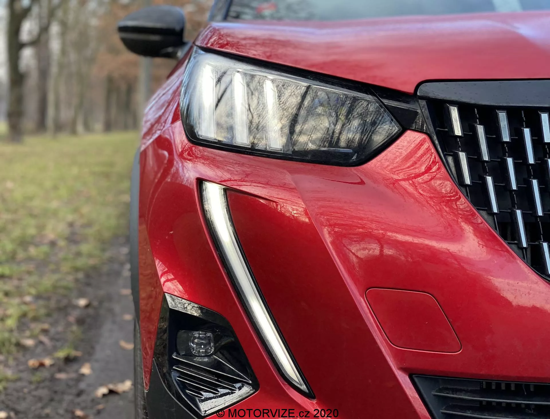 Zbliżenie z niskiego kąta na przednią lewą część Peugeot 2008 2020 z błyszczącym czerwonym wykończeniem. Obraz podkreśla elegancką, kątową lampę LED, ostrą konstrukcję wlotu powietrza i rzeźbione linie, odzwierciedlając zaawansowaną technologię oświetlenia i aerodynamiczną estetykę.