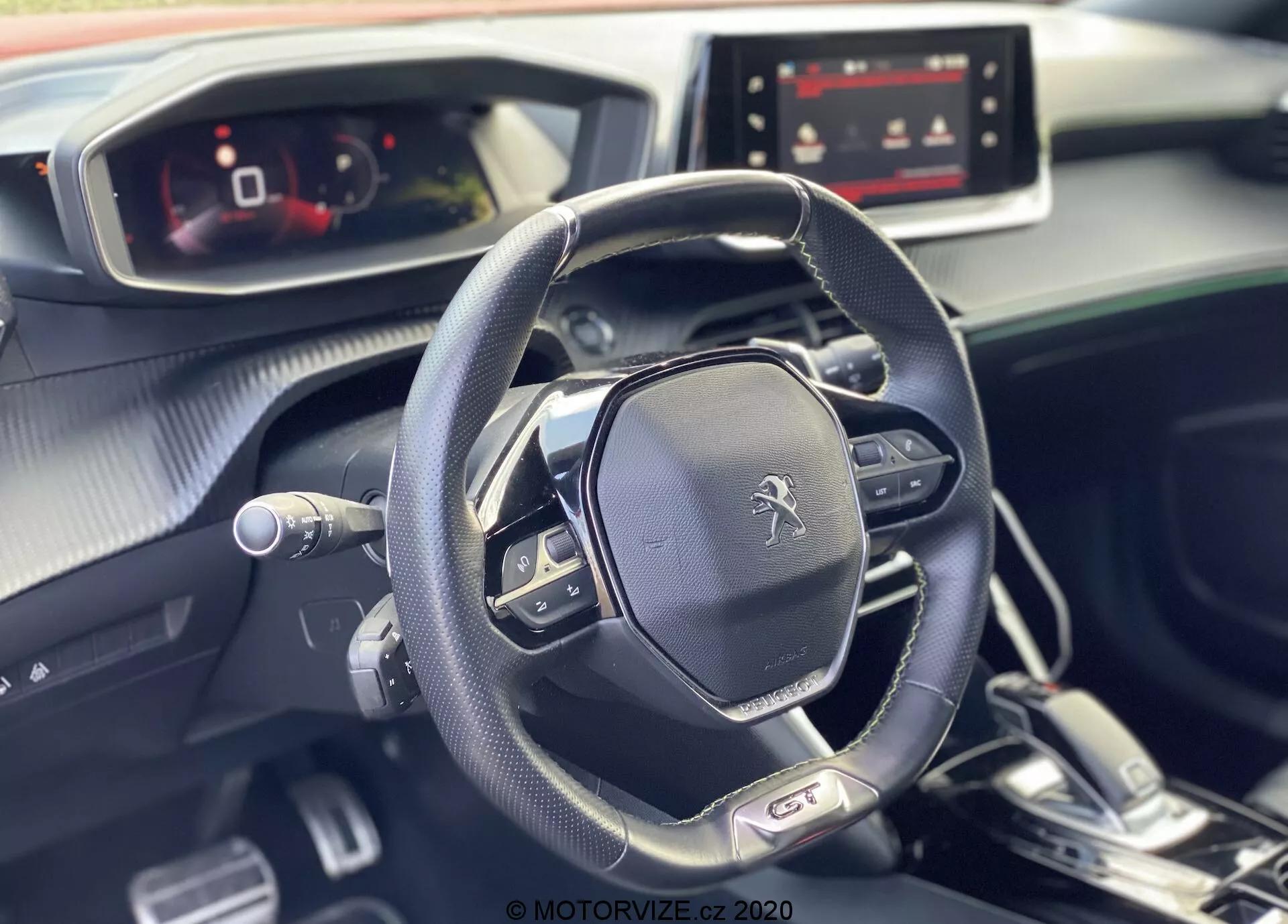 Obraz przedstawia nowoczesne i eleganckie wnętrze Peugeot 2008 2020, podkreślając wielofunkcyjną kierownicę i w pełni cyfrowy zestaw wskaźników z perspektywy pasażera. Na kierownicy wyraźnie widoczny jest emblemat Peugeot, który zawiera elementy sterujące mediami, ustawienia tempomatu i manetki, co sugeruje automatyczną skrzynią biegów z opcjami ręcznej zmiany biegów. Deska rozdzielcza, zaprojektowana z połączeniem materiałów miękkich w dotyku i metalicznych akcentów, zawiera włączony ekran multimedialny, wskazujący na różne opcje łączności i nawigacji. Naturalne światło podkreśla nieskazitelną i przyjemną atmosferę we wnętrzu pojazdu.