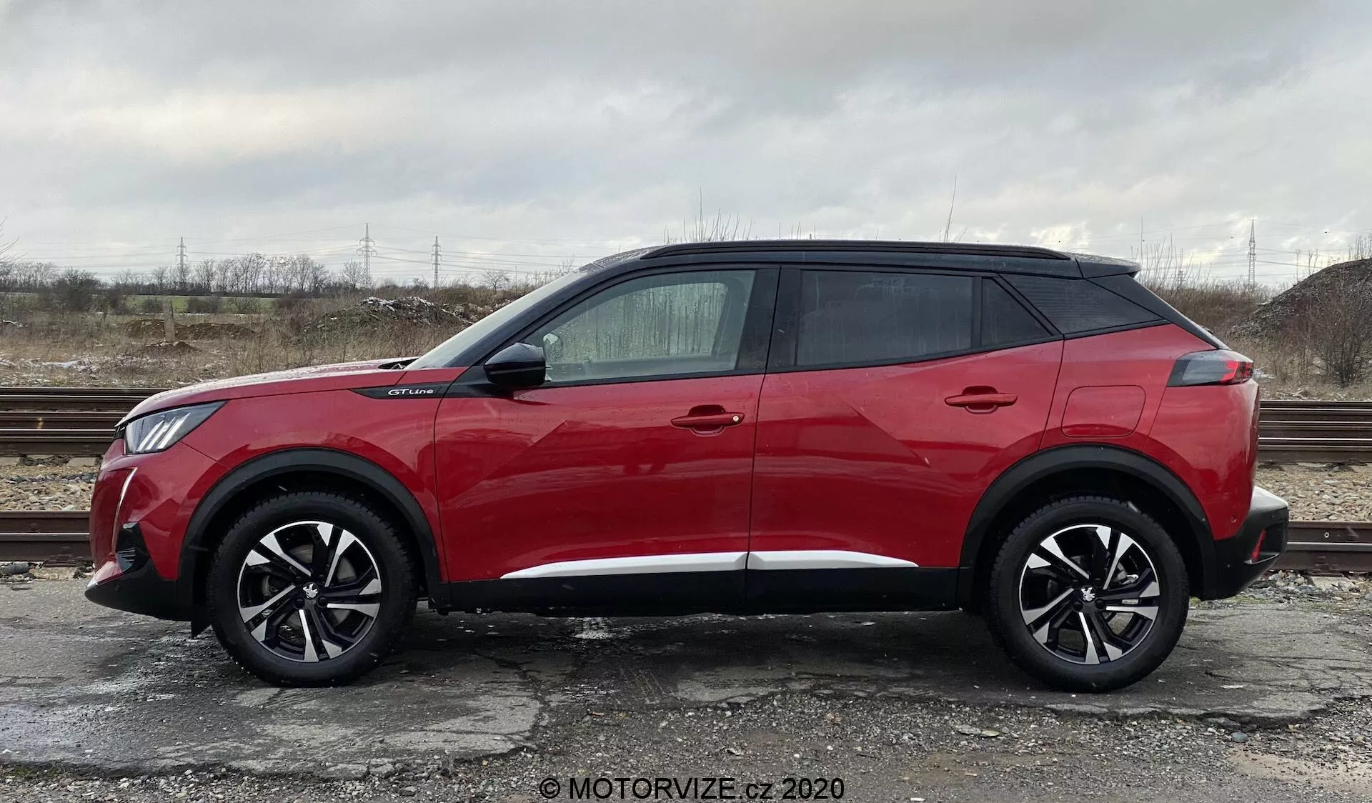 Profil boczny jasnoczerwonego Peugeot 2008 2020 SUV, model 1.2 PureTech 100 Active, zaparkowanego na żwirowej powierzchni z rzadką roślinnością i pochmurnym niebem w tle. Na zdjęciu widać pojazd z dużymi, czarnymi, wieloramiennymi felgami aluminiowymi, nowoczesnym eleganckim designem z wysoką linią boczną, czterema klamkami, przyciemnianymi szybami, wystającymi lusterkami bocznymi z wbudowanymi kierunkowskazami i unoszącym się dachem z czarnymi tylnymi słupkami. Linia ozdobna biegnąca wzdłuż dolnej części samochodu.