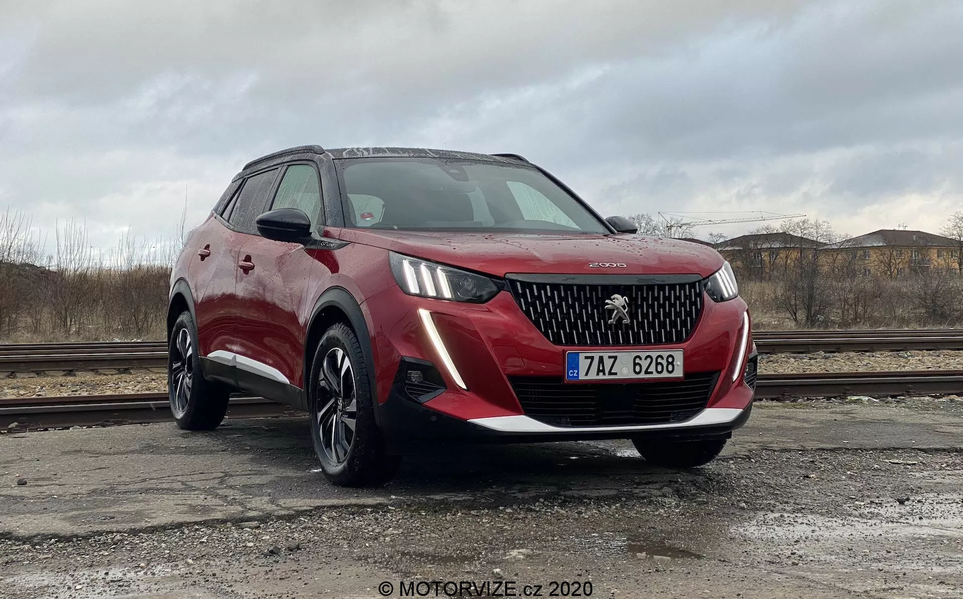 Czerwony SUV Peugeot 2008 2020 (Peugeot-2008-1-2-PureTech-100-Active) zaparkowany na zewnątrz pod pochmurnym niebem z suchym terenem i gruzami budowlanymi w tle. Pojazd został uchwycony z niskiego i bliskiego kąta przedniego trzech czwartych, pokazującego przednią kratkę z logo Peugeot, eleganckie reflektory LED, światła przeciwmgielne, wieloramienne felgi z lekkich stopów i metaliczne czerwone wykończenie lakiernicze z czarnymi plastikowymi elementami wokół nadkoli. Boczne lusterka, częściowo widoczne, wydają się być w kolorze nadwozia z wbudowanymi kierunkowskazami, a nachylona przednia szyba zapewnia dobrą widoczność kierowcy. Prawdopodobne funkcje to czujniki parkowania i kamery, co można wywnioskować z jego nowoczesnego designu.