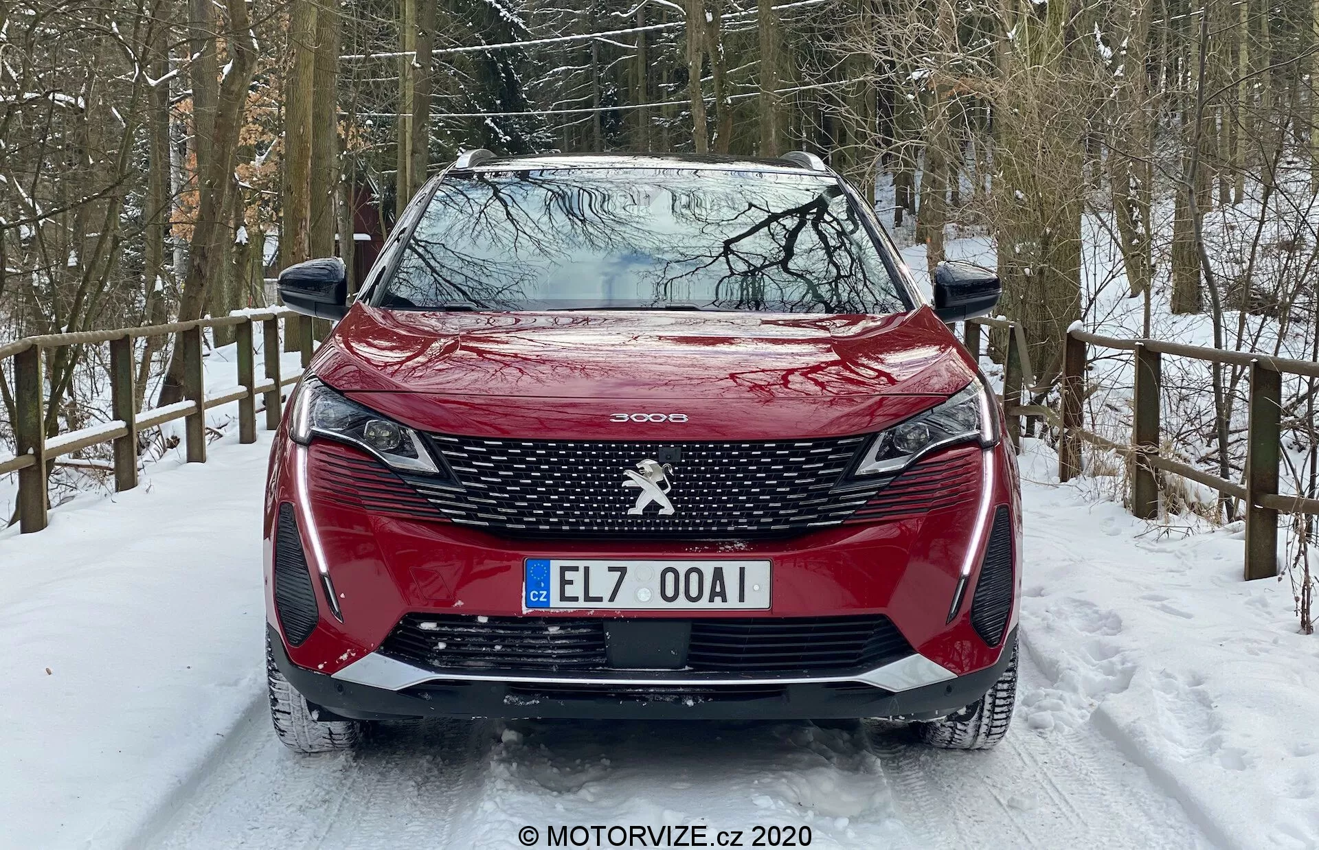 Widok z przodu czerwonego Peugeot 3008 2021 zaparkowanego na zaśnieżonej drodze, z charakterystycznym wzorem grilla, centralnym emblematem Peugeot, ostrymi światłami przednimi z dziennymi światłami w kształcie kłów, profilowaną maską, przednim zderzakiem z wbudowanymi czujnikami parkowania i zaśnieżonym otoczeniem leśnym.