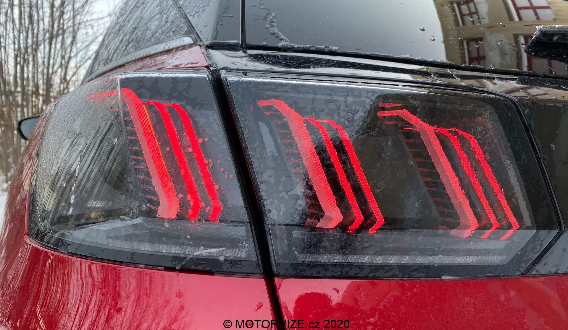 Zbliżenie tylnej części Peugeot 3008 z 2021 roku, podkreślające nowoczesny, pionowo podświetlany design tylnej lampy LED na błyszczącej czerwonej karoserii z kroplami wody, sugerując otoczenie na świeżym powietrzu podczas lub po deszczu; tylna lampa charakteryzuje się energooszczędną technologią i pełni funkcję wskaźników świateł hamowania i pozycyjnych.