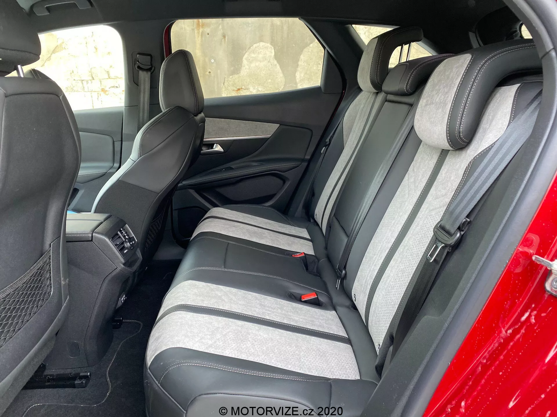 Widok wnętrza Peugeot 3008 z 2021 roku, koncentrujący się na tylnej części siedzeń, widziany z zewnątrz, pod kątem w stronę kierowcy. Tylne siedzenia mają dwu-tonową tkaninę w ciemnych i jasnych odcieniach szarości z teksturowanym wzorem pasków. Widoczne są dopasowane podniesione zagłówki. Część przednich siedzeń i konsoli środkowej jest również widoczna. Tylne siedzenia są wyposażone w trzypunktowe pasy bezpieczeństwa i podłokietnik z wbudowanymi uchwytami na napoje. Panele drzwi odpowiadają kolorom siedzeń, mają kratki głośników i sekcje sterowania energią.
