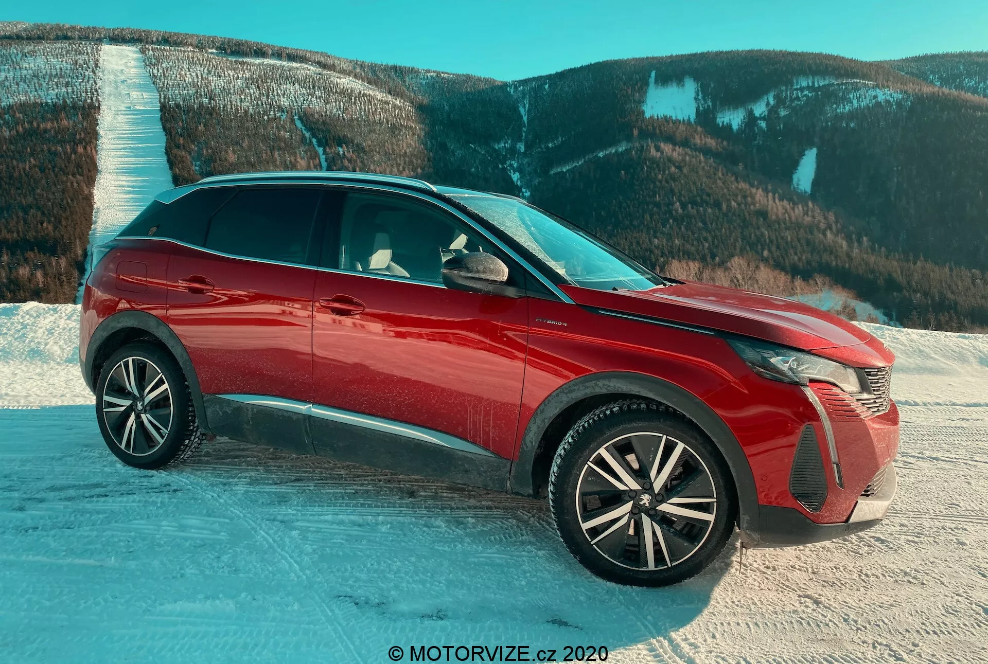SUV Peugeot 3008 z 2021 roku w czerwonym kolorze, sfotografowany z widoku trzy czwarte od przodu na zaśnieżonej powierzchni z górskim tłem, podkreślający zaawansowane reflektory LED, skomplikowane koła aluminiowe, błyszczące wykończenie, aktywne kratki chłodnicy oraz lusterka boczne z wbudowanymi kierunkowskazami, podkreślający cechy projektowe i przydatność do trudnych warunków.