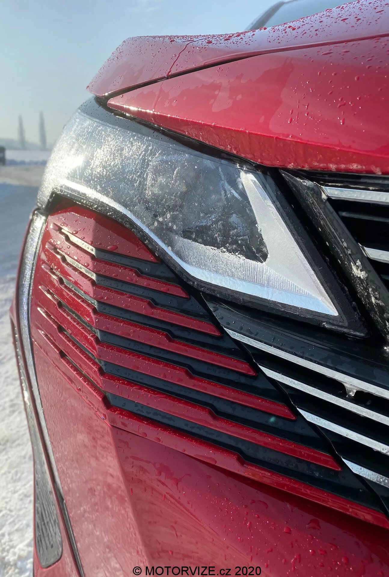 Zbliżenie na przednią część Peugeot 3008 2021, koncentrując się na nowoczesnym reflektorze LED i atrapie chłodnicy z pionowymi listwami, w metalicznym czerwonym kolorze pokrytym kroplami wody, sugerującymi niedawny deszcz lub szron. Śnieżne tło sugeruje zimowe warunki lub pielęgnację samochodu.