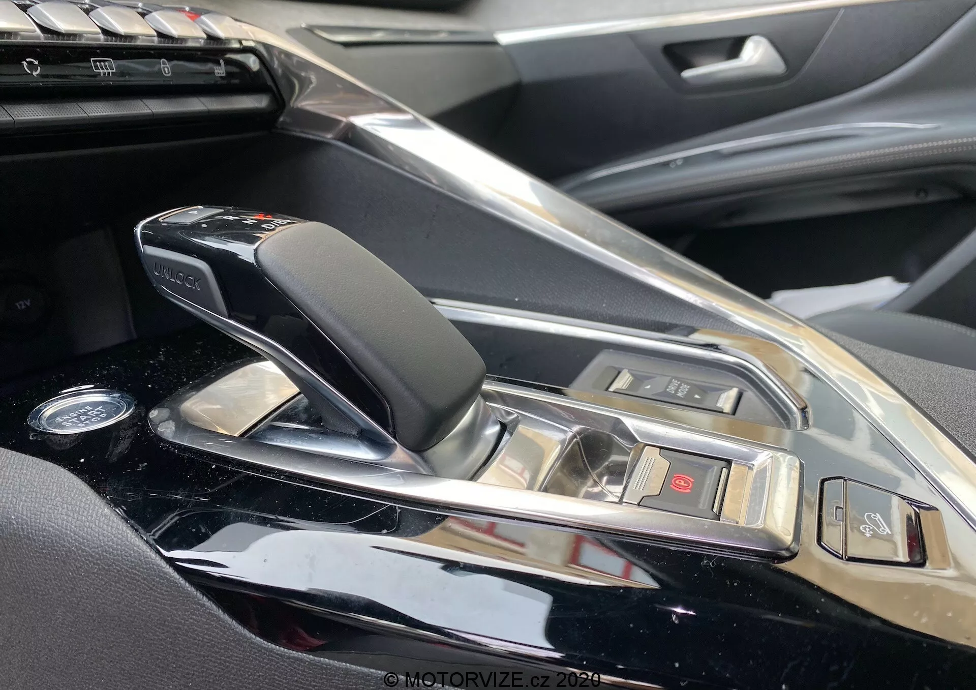 Zbliżenie na konsolę centralną i obszar dźwigni zmiany biegów w samochodzie Peugeot 3008 z 2021 roku, widok z perspektywy pasażera. Prezentuje nowoczesną automatyczną skrzynię biegów z eleganckim wzornictwem, skórzanym i chromowanymi elementami, otoczoną połyskującym wykończeniem konsoli. Widoczne przyciski do różnych funkcji pojazdu i fragment systemu multimedialnego wskazują na luksusowe wnętrze samochodu z wysokiej jakości materiałami i zaawansowanymi funkcjami technologicznymi.