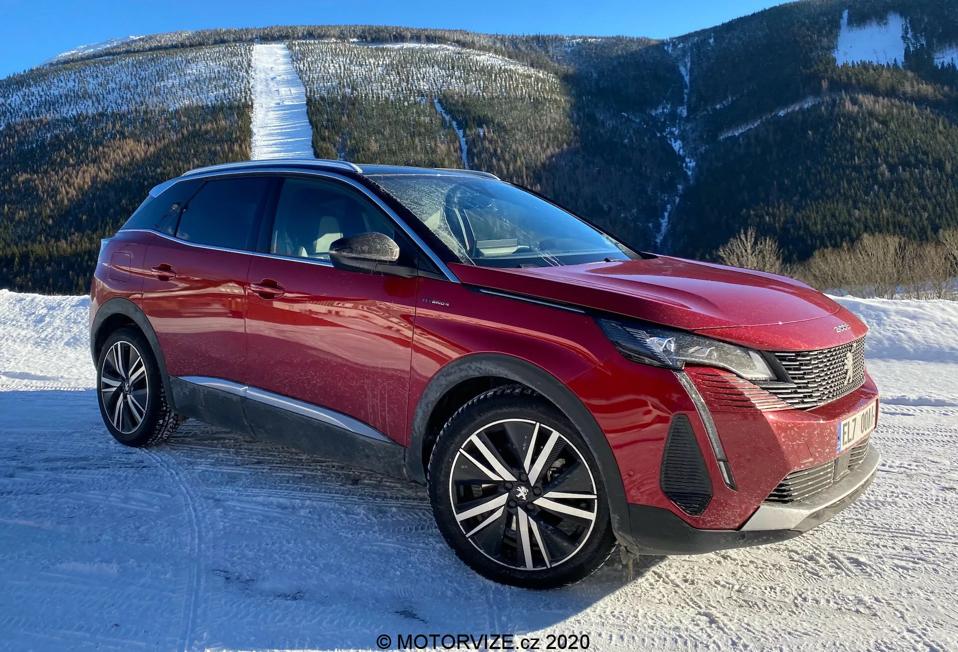 Obraz nowoczesnego samochodu, prawdopodobnie Peugeot 3008 z 2021 roku, zrobiony z przedniej trzykwarowej perspektywy. Kąt ukazuje charakterystyczny design reflektorów LED samochodu, kratkę w kształcie grzebienia i emblemat z przodu. Przedni zderzak ma sekcje wlotu powietrza oraz obudowy świateł przeciwmgielnych. Maska ma rzeźbione linie, które przyczyniają się do aerodynamiki, a szeroka szyba przednia sugeruje dobrą widoczność do przodu. Felgi o wieloramiennym wzorze sugerują bardziej sportowy wygląd i wyższy poziom wyposażenia. Smukła linia dachu oraz dynamiczne linie nadwozia sugerują crossover lub SUV. Widoczne są także lusterka boczne z wbudowanymi kierunkowskazami oraz elementy ozdobne po bokach. Scena przedstawia zaśnieżoną drogę z górskim tłem, co sugeruje zdolność pojazdu do radzenia sobie z różnymi terenami.