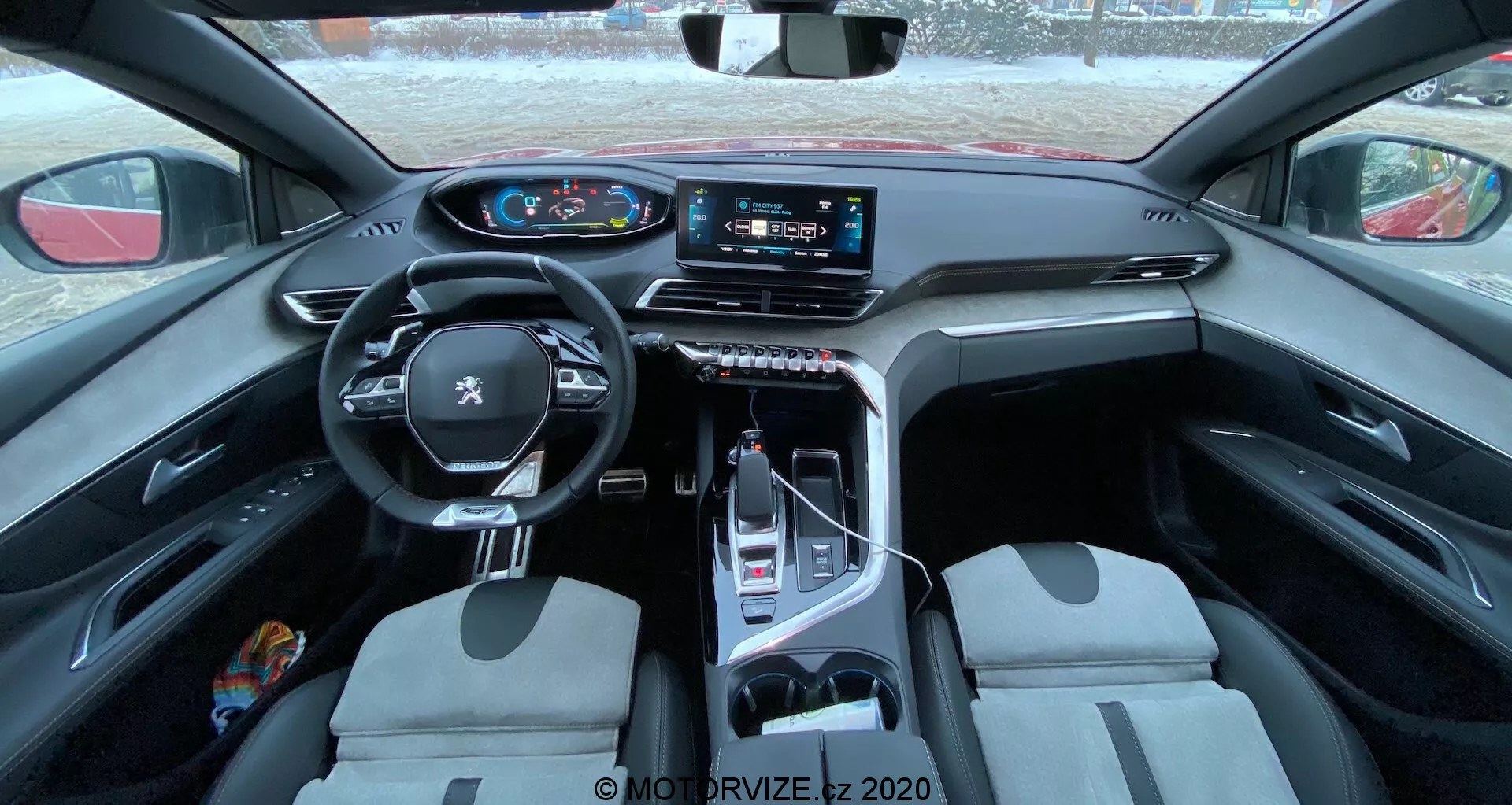 Widok wnętrza Peugeot 3008 z 2021 roku, pokazujący wielofunkcyjną, skórzaną kierownicę z zamontowanymi przyciskami, cyfrowy zestaw wskaźników, duży dotykowy system informacyjno-rozrywkowy na konsoli centralnej, elektroniczny selektor biegów, przednie fotele w dwukolorowej konfiguracji oraz mieszankę praktycznych i luksusowych materiałów. Zdjęcie wykonane z tylnego siedzenia, zapewniające rozległy widok na kabinę, podkreślającą design i cechy technologiczne.