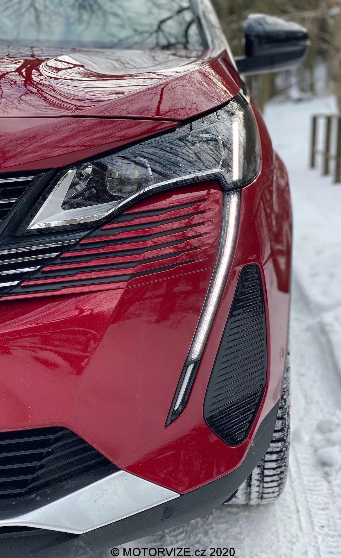 Zbliżenie nisko kąta na nowoczesny czerwony Peugeot 3008 2021. Skupienie na przedniej-lewej ćwiartce ukazuje eleganckie kątowe reflektory LED, dużą agresywną kratę, wyrazisty projekt kratek wentylacyjnych, błyszczący lakier i ostro konturowane panele nadwozia. Lusterko boczne z wbudowanymi kierunkowskazami, a na scenie widoczna jest zaśnieżona ziemia sugerująca zimowe warunki. Widoczny znak wodny MOTORVIZE.cz.