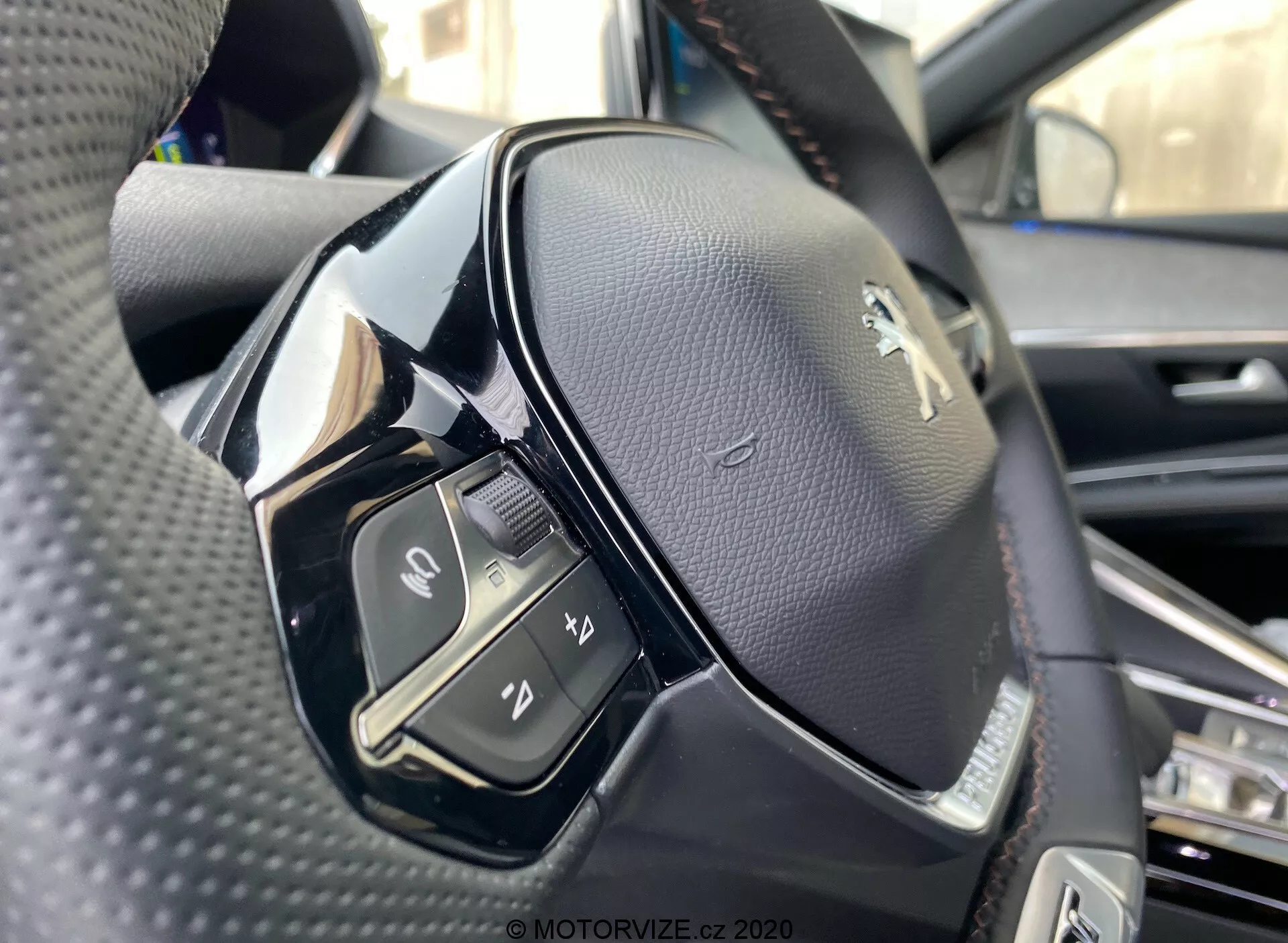 Zbliżenie widoku kierownicy z perspektywy siedzenia kierowcy w Peugeot 3008 2021. Obraz uwydatnia lewą stronę kierownicy, wykonany z nieco niższego kąta, ukazując szczegóły lewej strony przycisków sterowania i logo Peugeot. Kierownica jest pokryta czarną perforowaną skórą z mieszanką materiałów matowych i błyszczących. Widoczne przyciski obejmują przełącznik, przyciski do funkcji multimedialnych i komunikacyjnych, takich jak akceptowanie lub kończenie połączeń, aktywację sterowania głosowego i inną niezidentyfikowaną funkcję. Ostrość jest wysoka, pokazuje szwy na skórze i błyszczące wykończenie na niektórych plastikowych częściach, prawdopodobnie przyciski dotykowe.
