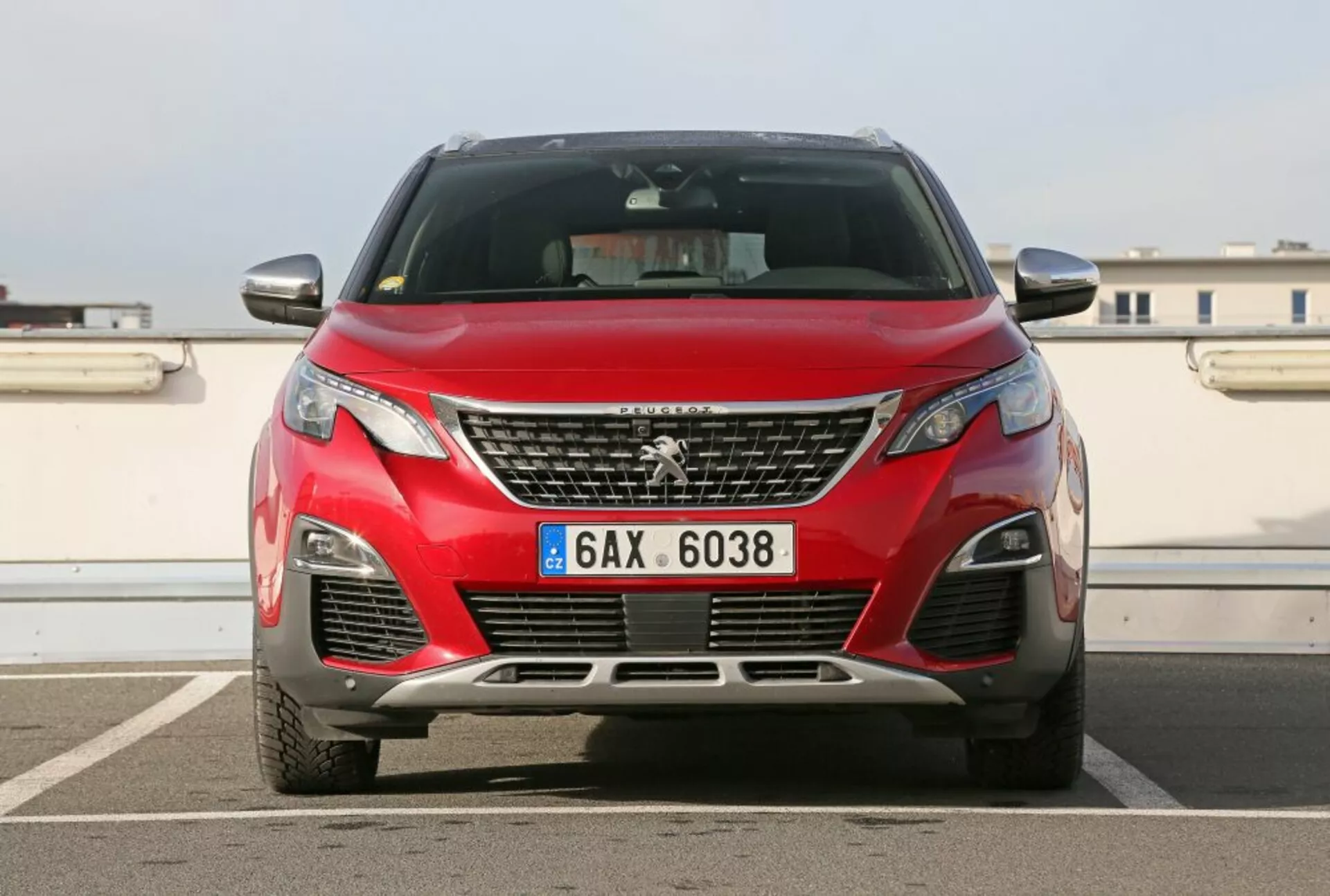 Przedni widok czerwonego Peugeot 3008 z 2016 roku prezentującego emblemat na grillu, kątowe reflektory, dolne wloty powietrza z lampami przeciwmgielnymi, sugestię konturów maski na tle miejskiego krajobrazu z jasnym niebem