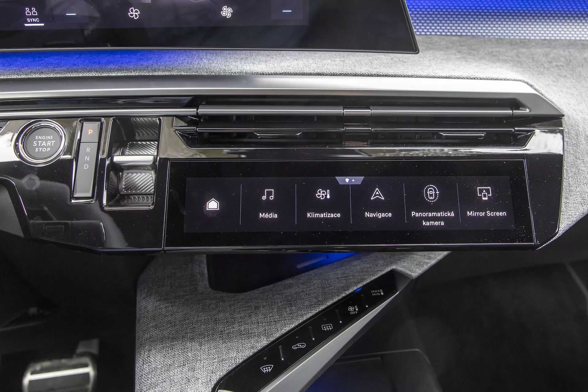 Zbliżenie konsoli środkowej i deski rozdzielczej Peugeot 3008 2024 (e-3008) z nowoczesnym systemem infotainment z ekranem dotykowym, który posiada ikony dla mediów, klimatyzacji, nawigacji GPS, kamery panoramicznej i lustrzanego odbicia smartfona. Obraz pokazuje również czarny błyszczący wykończenie, teksturę przypominającą tkaninę poniżej konsoli i przycisk START STOP, podkreślając zaawansowaną technologię i elegancki design wnętrza pojazdu.