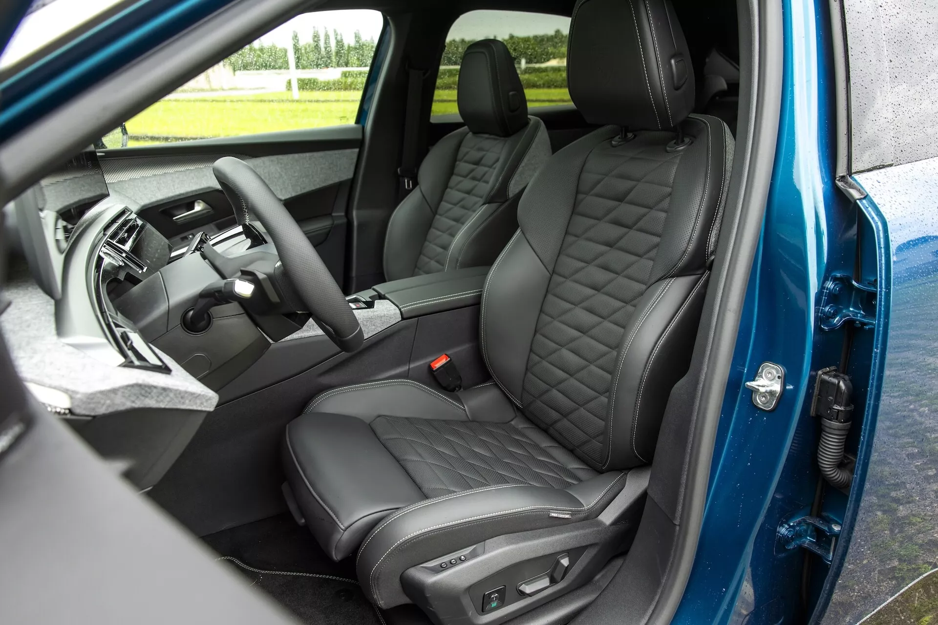 Szczegółowy widok wnętrza samochodu Peugeot 3008 2024 (e-3008) z lekkiego podwyższenia po stronie pasażera. Otwarte przednie drzwi pasażera odsłaniają pikowane skórzane fotele z diamentowym przeszyciem, premiumową konsolę środkową, ekran infotainment, wielofunkcyjną kierownicę po lewej stronie i elektryczne sterowanie szybami z ustawieniami pamięci na drzwiach. Ciemne i luksusowe materiały podkreślają nowoczesne skoncentrowanie samochodu na komforcie i technologii.