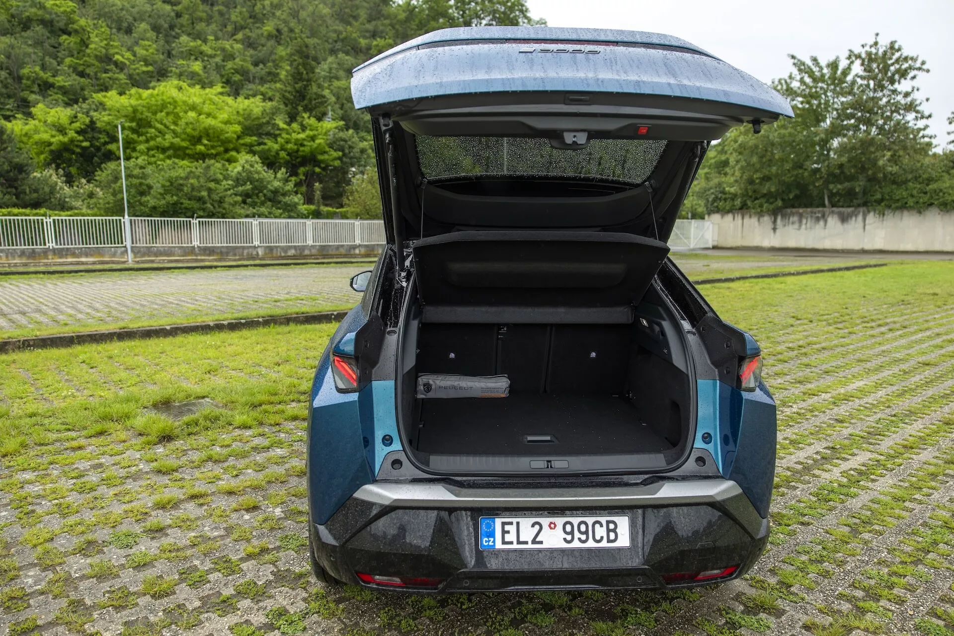 Tylny widok hatchbacka Peugeot 3008 2024 e-3008, uchwycony z niskiego kąta w ciągu dnia na zewnątrz. Tylny bagażnik jest otwarty, ukazując przestronną przestrzeń ładunkową. Widoczna tablica rejestracyjna z oznaczeniem 'EL', eleganckie tylne światła LED, tylne odblaski i czujniki parkingowe. Dwukolorowe malowanie z ciemnoniebieskim korpusem głównym i czarną lub ciemnoszarą dolną sekcją. Tylna szyba zawiera wycieraczkę, podkreślając praktyczność i nowoczesny styl, uzupełniony ekologicznym rozwiązaniem parkingowym z trawą między betonowymi blokami.