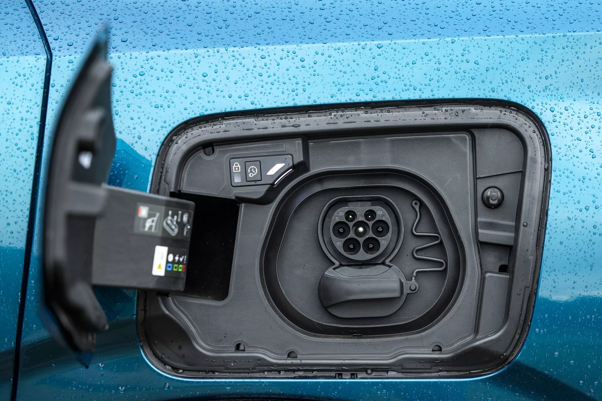 Zbliżenie obszaru portu ładowania w elektrycznym pojeździe Peugeot 3008 2024 (e-3008). Zdjęcie wykonane pod niewielkim kątem z góry i z boku pokazuje otwartą pokrywę portu ładowania, odsłaniającą gniazdo z wieloma połączeniami stykowymi. Wtyczka jest częściowo widoczna na pierwszym planie po lewej stronie, z przewodem wychodzącym z kadru, co wskazuje, że jest gotowa do włożenia lub dopiero została usunięta. Krople wody na niebieskiej karoserii sugerują ostatni deszcz lub mokre środowisko, podkreślając odporną na warunki atmosferyczne konstrukcję portu.