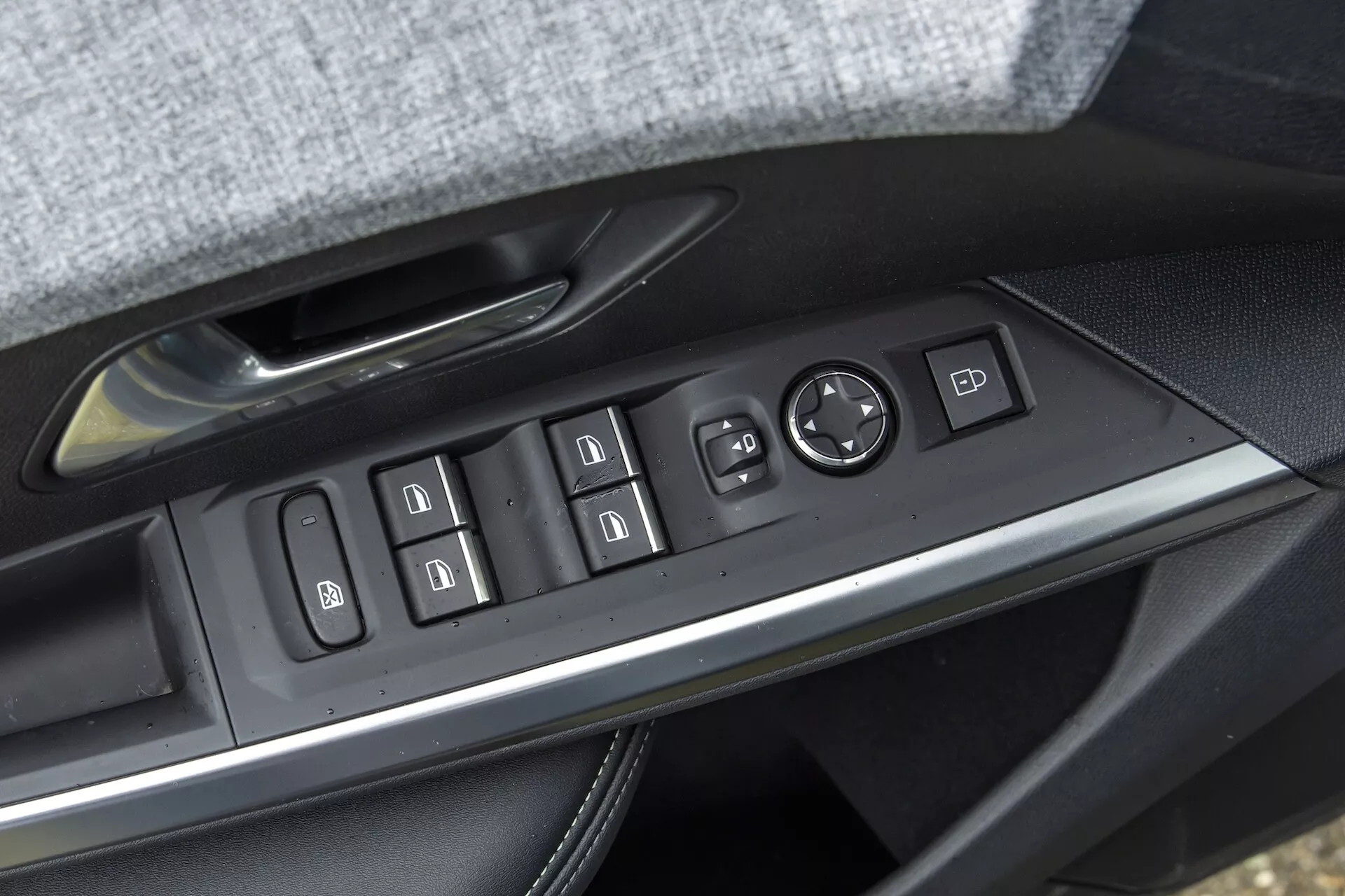 Zbliżenie panelu sterowania drzwiami kierowcy w modelu 2024 Peugeot e-3008. Funkcje obejmują sterowanie wszystkimi czterema oknami z funkcją automatyczną, blokadę okien, regulację i składanie lusterek bocznych, przyciski blokady i odblokowywania drzwi oraz ustawienia pamięci fotela. Panel ma podświetlane ikony i jest wykończony szczotkowanym metalem, osadzony w tapicerowanym podłokietniku, być może ze skóry lub podobnego materiału.