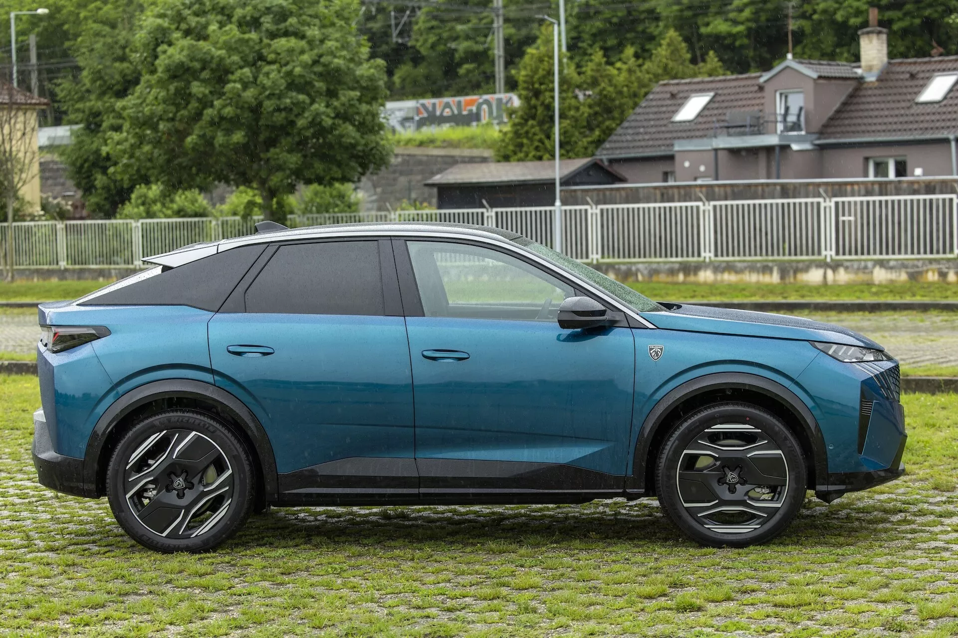 Profil boczny eleganckiego i nowoczesnego elektrycznego SUV-a, prawdopodobnie Peugeot 3008 2024 (e-3008), uchwycony w świetle dziennym. Głęboko niebieski pojazd z błyszczącym wykończeniem ma kontrastujący czarny dach, czarne osłony nadkoli, płaskie klamki drzwi i pięć drzwi, co wskazuje na praktyczny, rodzinny design. Pojazd stoi na trawiastym terenie z ogrodzeniem i budynkami miejskimi lub podmiejskimi w tle, wyposażony w wieloszprychowe, czarno-srebrne felgi aluminiowe i w pełni przyciemniane szkła, co zwiększa prywatność i chroni przed promieniowaniem UV. Lusterka zintegrowane z kierunkowskazami poprawiają bezpieczeństwo, wskazując na zaawansowane funkcje technologiczne.