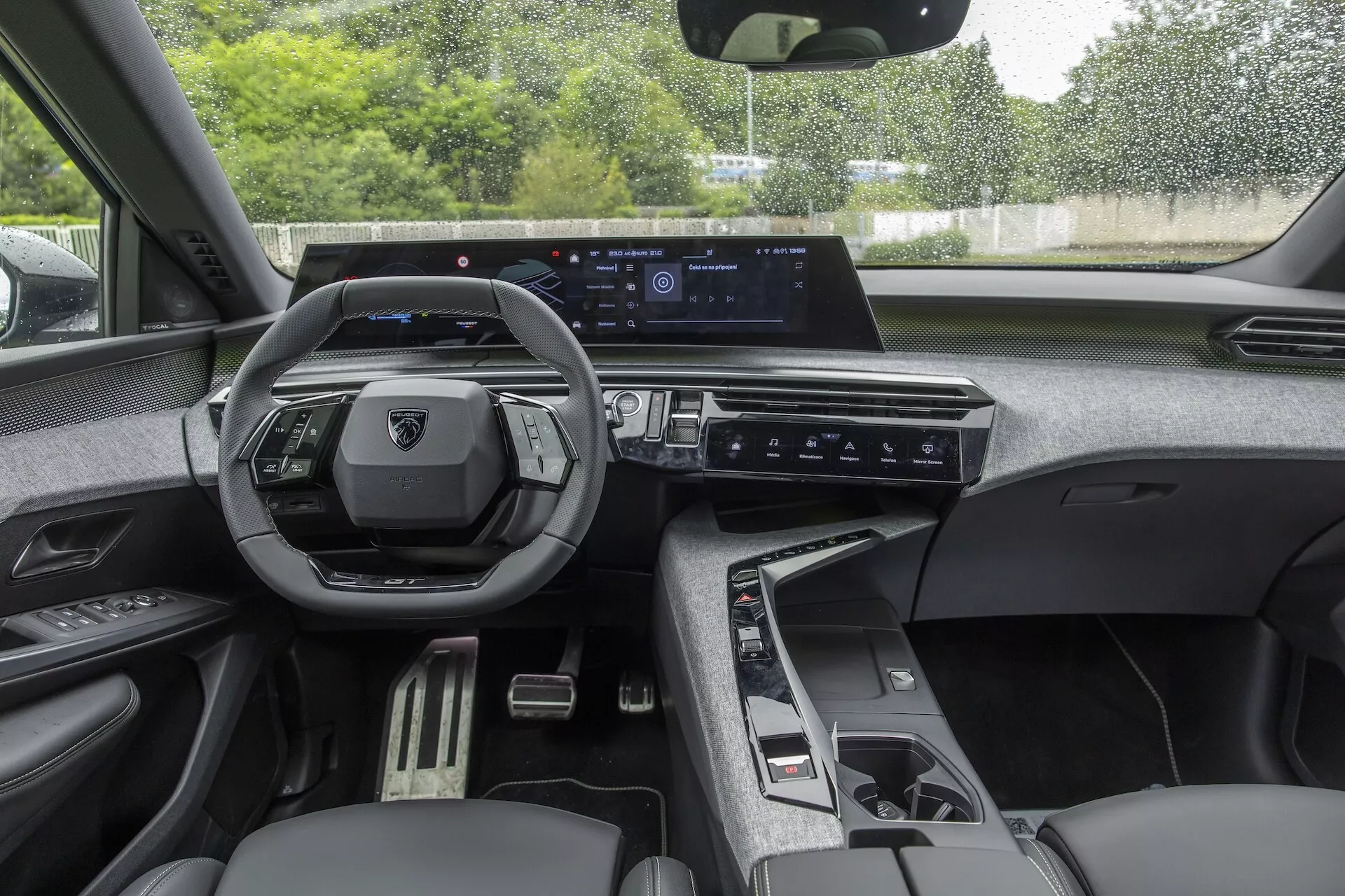 Widok wnętrza z siedzenia kierowcy Peugeot 3008 (e-3008) 2024 z nowoczesną, wielofunkcyjną kierownicą o płaskim dole, cyfrowym zestawem wskaźników, dużym poziomym ekranem dotykowym systemu infotainment, eleganckim panelem klimatyzacji, automatyczną elektroniczną dźwignią zmiany biegów oraz ciemnym tapicerowaniem siedzeń z szczegółowymi przeszyciami. Funkcje obejmują sterowanie dźwiękiem, telefonem, tempomatem, konfigurowalny wyświetlacz cyfrowy, nawigację, kompatybilność z Apple CarPlay/Android Auto, minimalne dotykowe sterowanie klimatyzacją oraz szczegóły wnętrza premium.