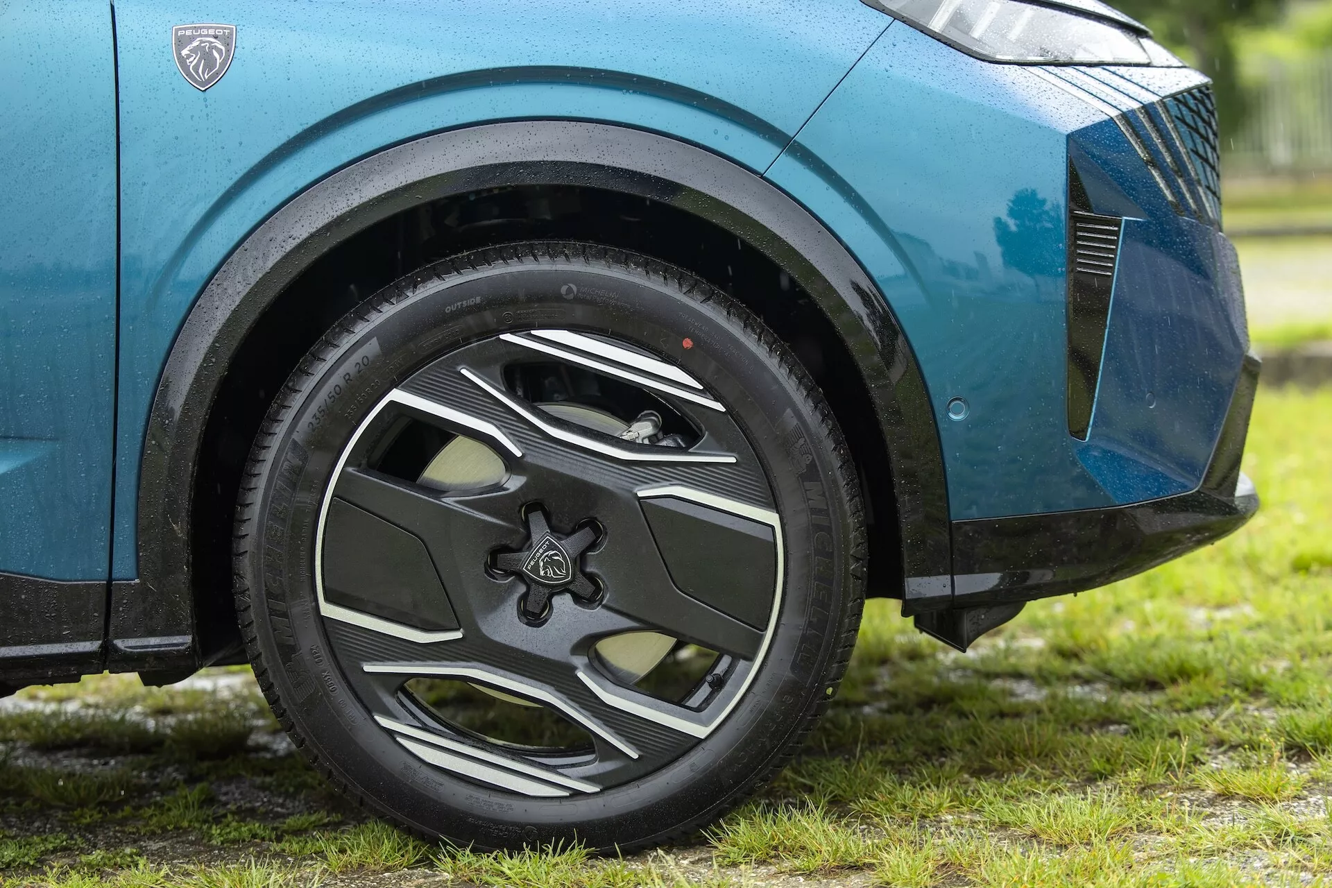 Zbliżenie na przód koła samochodu Peugeot 3008 2024 (e-3008) z niskiego kąta, podkreślające jego skomplikowany design z wieloma szprychami i dynamiczną przednią częścią nadkola. Samochód jest zaparkowany na trawie, co sugeruje naturalne lub zewnętrzne warunki. Metaliczny niebieski lakier lekko błyszczy, wskazując na czystą powierzchnię lub niedawny deszcz. Widoczny jest wzór bieżnika opony, część przedniego zderzaka i dolna kratka. Sceneria sugeruje naturalne światło dzienne.