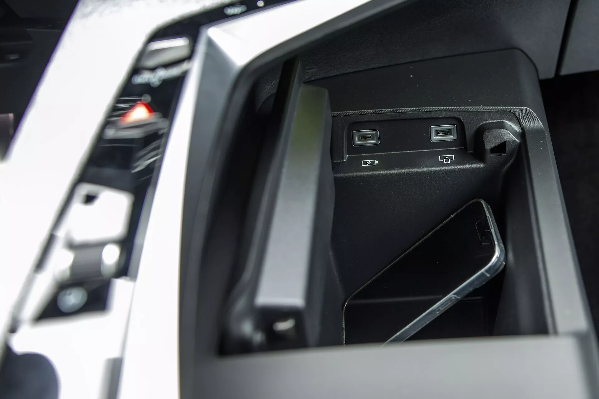 Zbliżenie na schowek w konsoli centralnej Peugeot 3008 2024 (e-3008) prezentującej opcje łączności, z dwoma portami USB, gniazdem na kartę SD i telefonem komórkowym umieszczonym w wysokiej jakości, błyszczącym czarnym wnętrzu schowka.