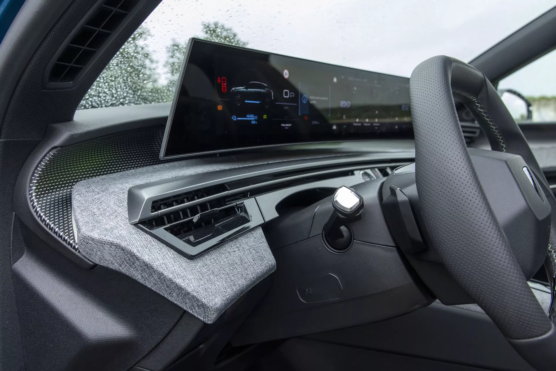 Widok wnętrza Peugeot 3008 (e-3008) 2024 z perspektywy kierowcy, skupiający się na desce rozdzielczej. Częściowo widoczna jest kierownica z zamontowanymi kontrolkami, z logo producenta pośrodku. Szeroki poziomy ekran infotainmentu dominuje na desce rozdzielczej, oferując diagnostykę pojazdu, nawigację i kontrole mediów. Poniżej ekranu znajdują się minimalistyczne centralne wloty powietrza z wykończeniem przypominającym włókno węglowe, sugerujące luksusowe wnętrze. Brak tradycyjnych analogowych wskaźników wskazuje na całkowicie cyfrową deskę rozdzielczą, podnosząc jej technologiczny urok. Luksusowe tekstury i materiały nadają wnętrzu sportowy, a zarazem ekskluzywny charakter.