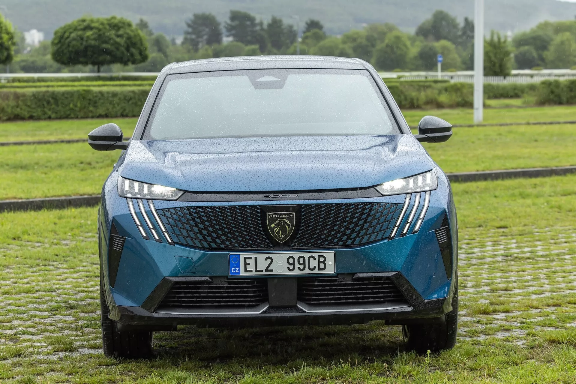 Widok z przodu nowoczesnego samochodu (Peugeot 3008 2024) e-3008, kąt prosty pokazujący osłonę chłodnicy z dużym emblematem, wzór geometryczny, reflektory LED z charakterystycznym podpisem świetlnym oraz konturowaną maskę. Widoczne cechy obejmują obudowy świateł przeciwmgielnych, aerodynamiczny dolny wlot powietrza, lusterka boczne z zintegrowanymi kierunkowskazami, wycieraczki i tablicę rejestracyjną zamontowaną centralnie.