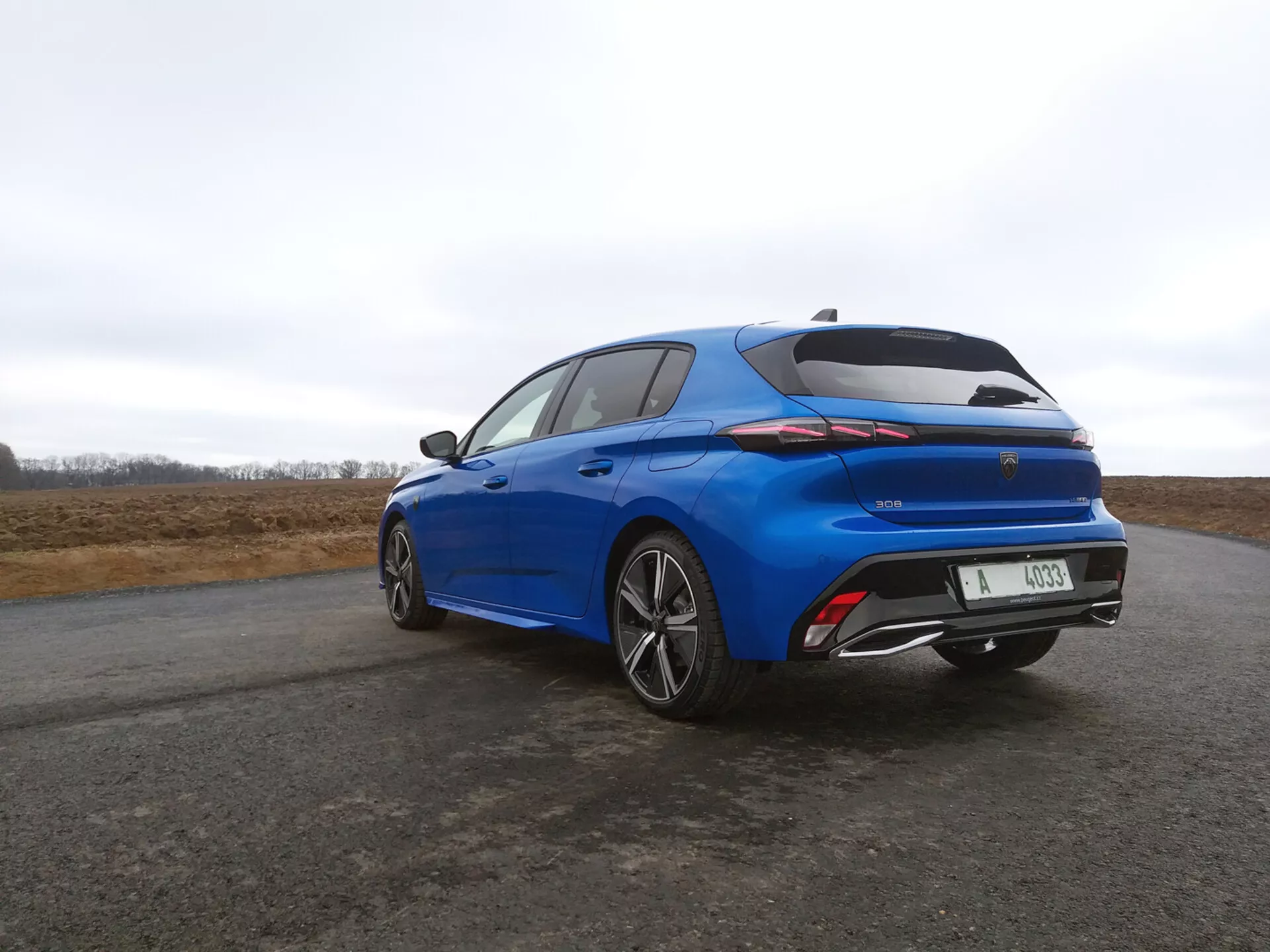 Hatchback Peugeot 308 z 2022 roku w żywym niebieskim kolorze, widok z tyłu pod kątem trzech czwartych, prezentujący smukłe tylne światła LED, zintegrowany spoiler i wieloramienne koła ze stopu na tle pochmurnego nieba.