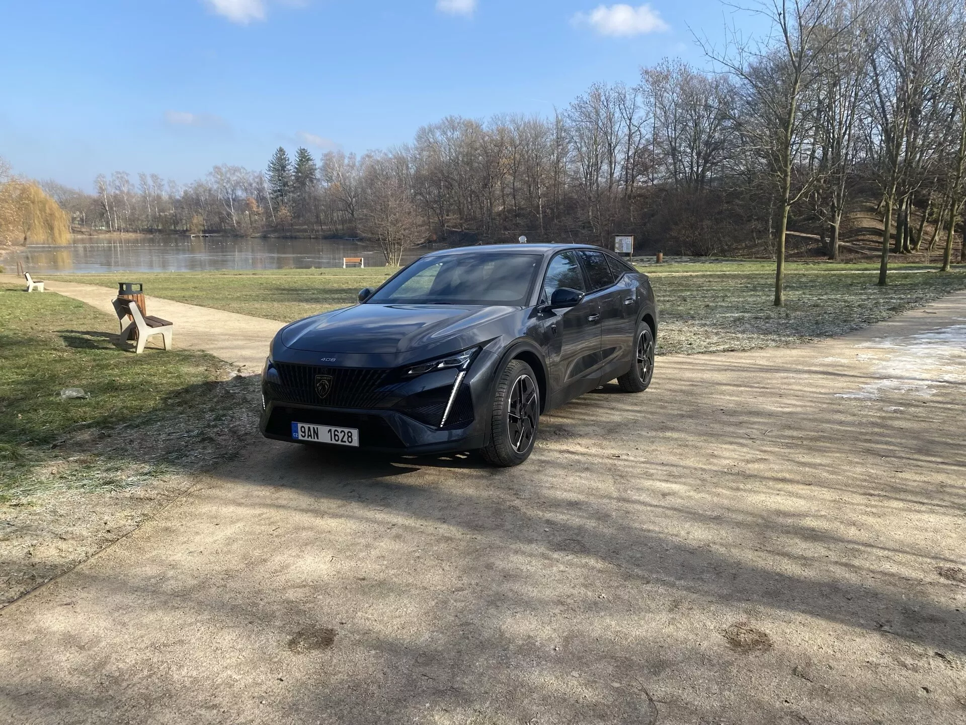 Peugeot 408 z 2023 roku zaparkowany w scenerii naturalnej podczas zimy lub wczesnej wiosny, prezentujący jego nowoczesny design crossover, reflektory LED, przedni system czujników, felgi aluminiowe i ekskluzywne wykończenie z perspektywy trzy czwarte z przodu pod światłem słonecznym z efektem flare obiektywu.