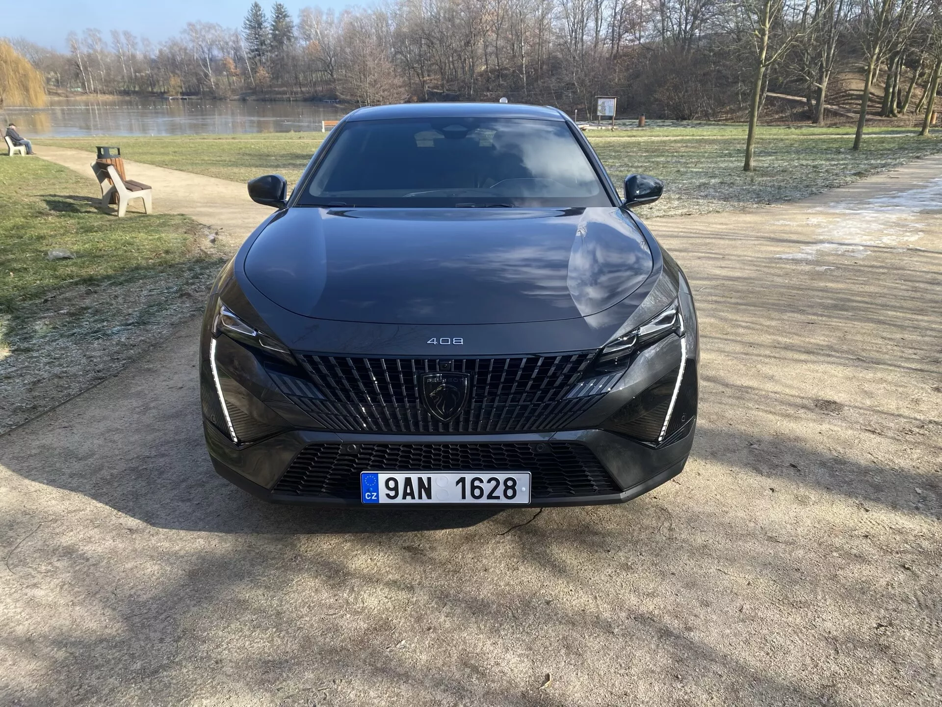 Przedni widok Peugeota 408 2023 zaparkowanego na żwirowej powierzchni z drzewami i zbiornikiem wodnym w tle, podkreślający jego dużą grill z wzorami w kształcie diamentów, eleganckie reflektory LED, rzeźbione linie na masce i zaawansowane funkcje wsparcia kierowcy