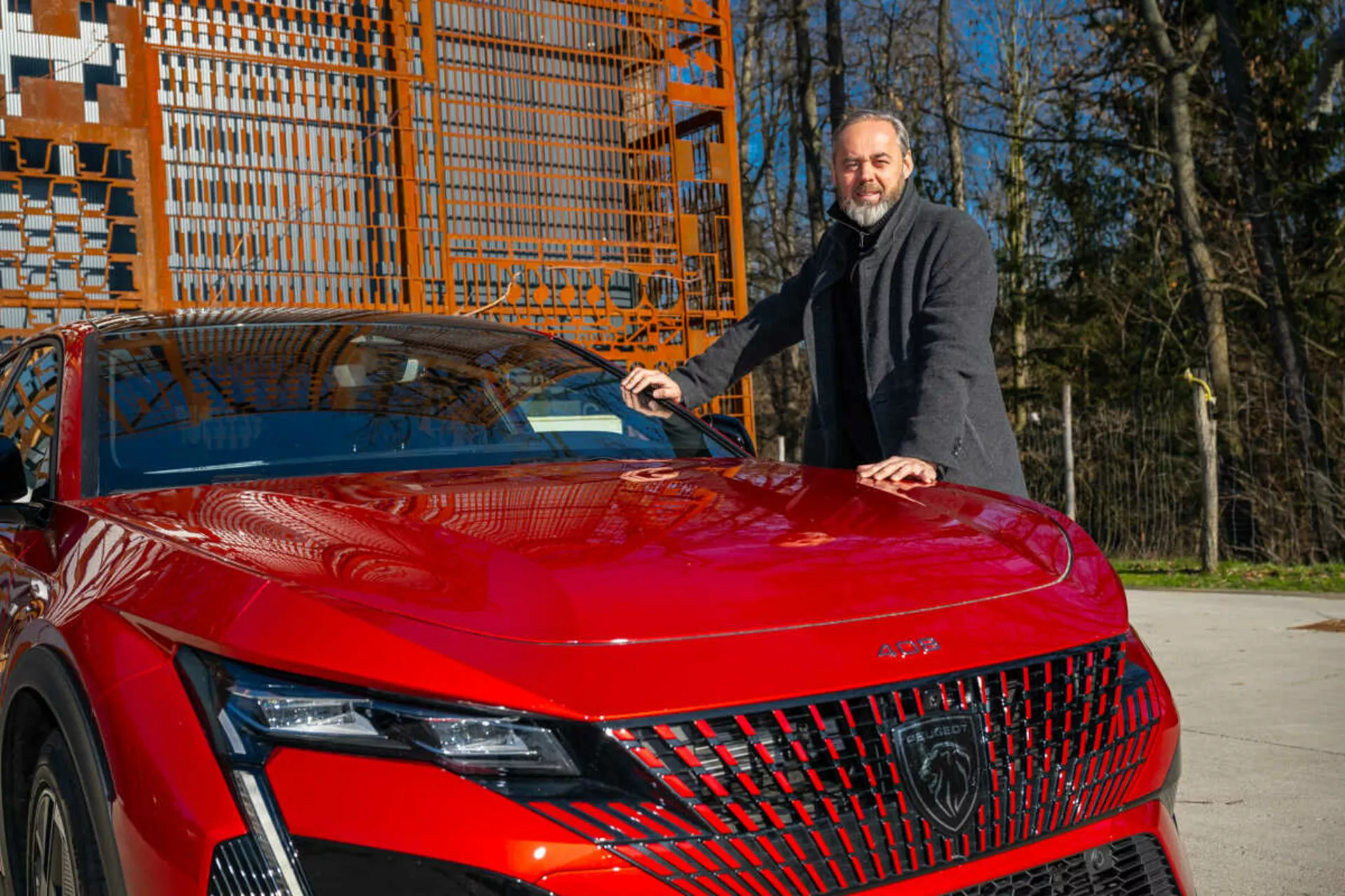 Czerwony model Peugeot 408 z 2023 roku prezentujący swój lewy przedni kwadrant w świetle dziennym, podkreślający szczegółowe reflektory LED, geometryczną kratę i sportową, aerodynamiczną maskę z mężczyzną w ciemnym płaszczu obok niego, który kładzie rękę na masce