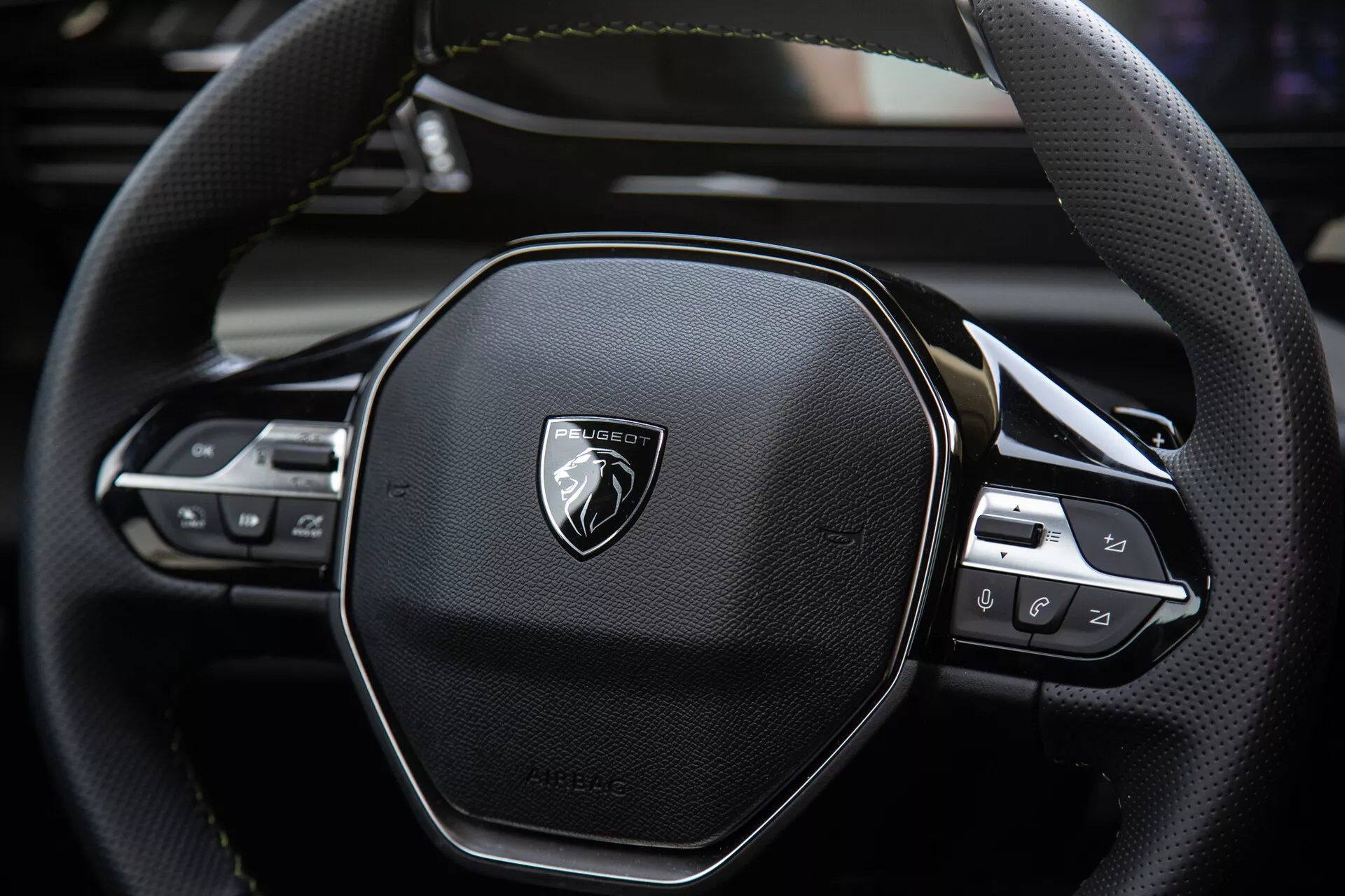 Zbliżenie na kierownicę Peugeot 408 2023 ukazujące ergonomiczny sześciokątny design, szwy z prawdziwej skóry, zintegrowane kontrole technologiczne dla audio i ustawień tempomatu, z emblematem na szczotkowanym metalu.