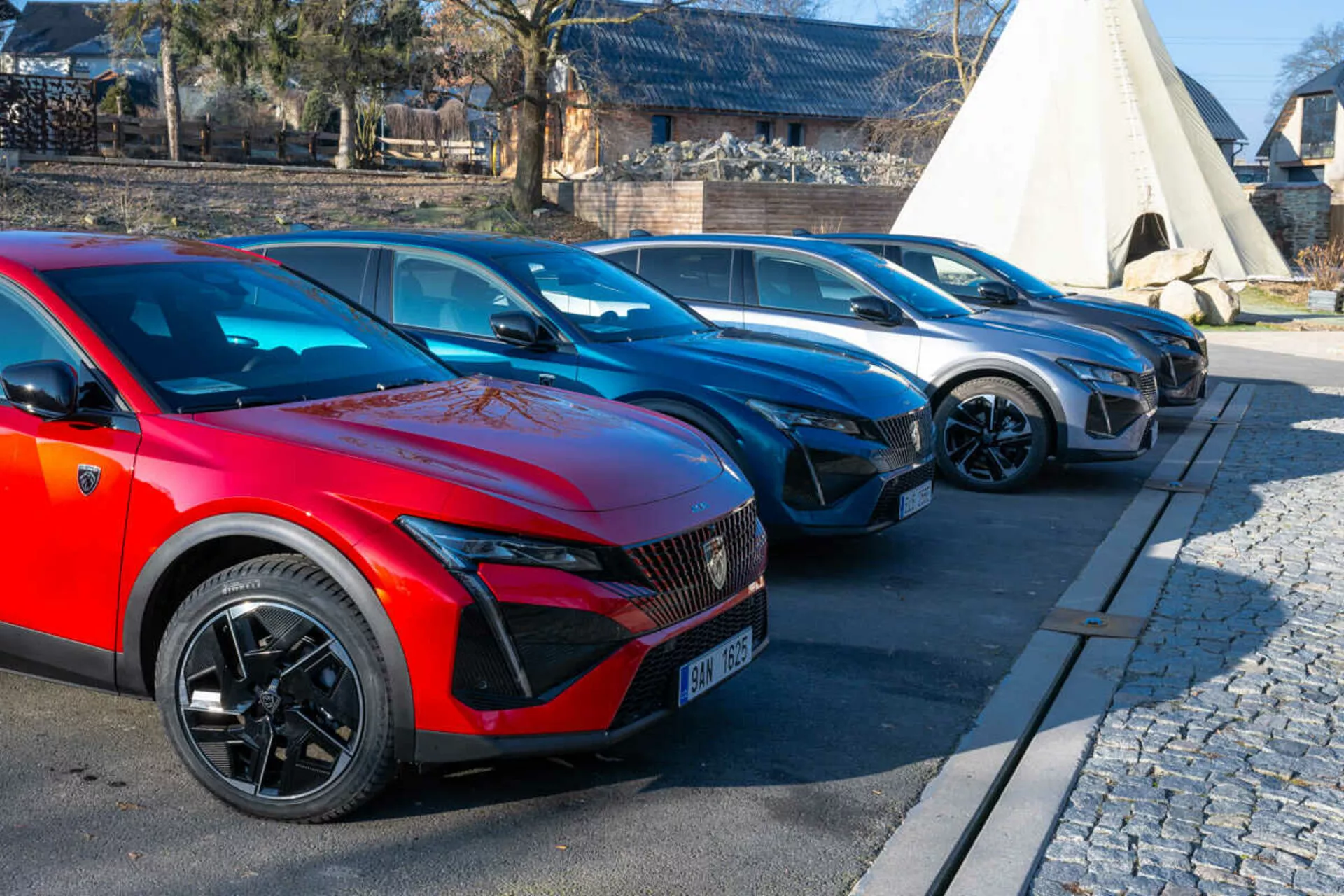 Lineup Peugeot 408 z 2023 roku z czerwonym samochodem na pierwszym planie z sportowymi kołami o wielu szprychach i światłami LED wskazującymi na modele elektryczne lub hybrydowe, zaparkowanym na wybrukowanym obszarze obok bruku
