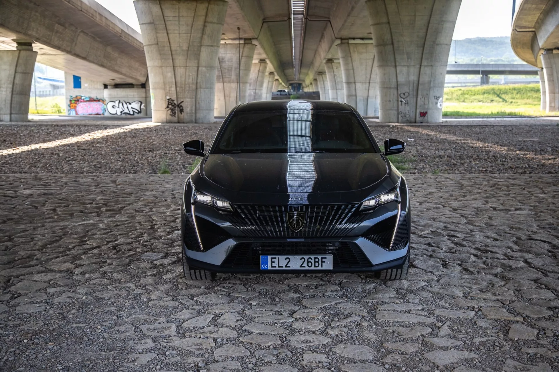 Peugeot 408 z 2023 roku w dynamicznej widoczności frontalnej podkreślającej agresywną postawę i sportowy design z detaliczną atrapą, smukłymi reflektorami LED i funkcjami aerodynamicznymi na wzorowanej nawierzchni pod miejskim mostem