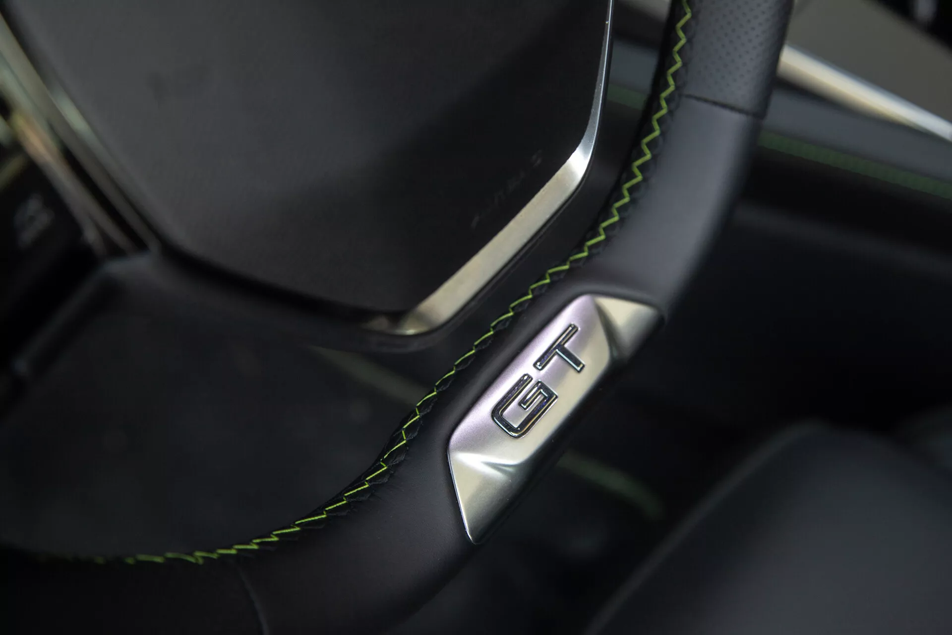 Zbliżenie na sekcję kierownicy i deski rozdzielczej Peugeot 408 2023, prezentująca czarną skórzaną kierownicę z zielonymi szwami i odznaką GT, podkreślającą luksus i sportowy charakter