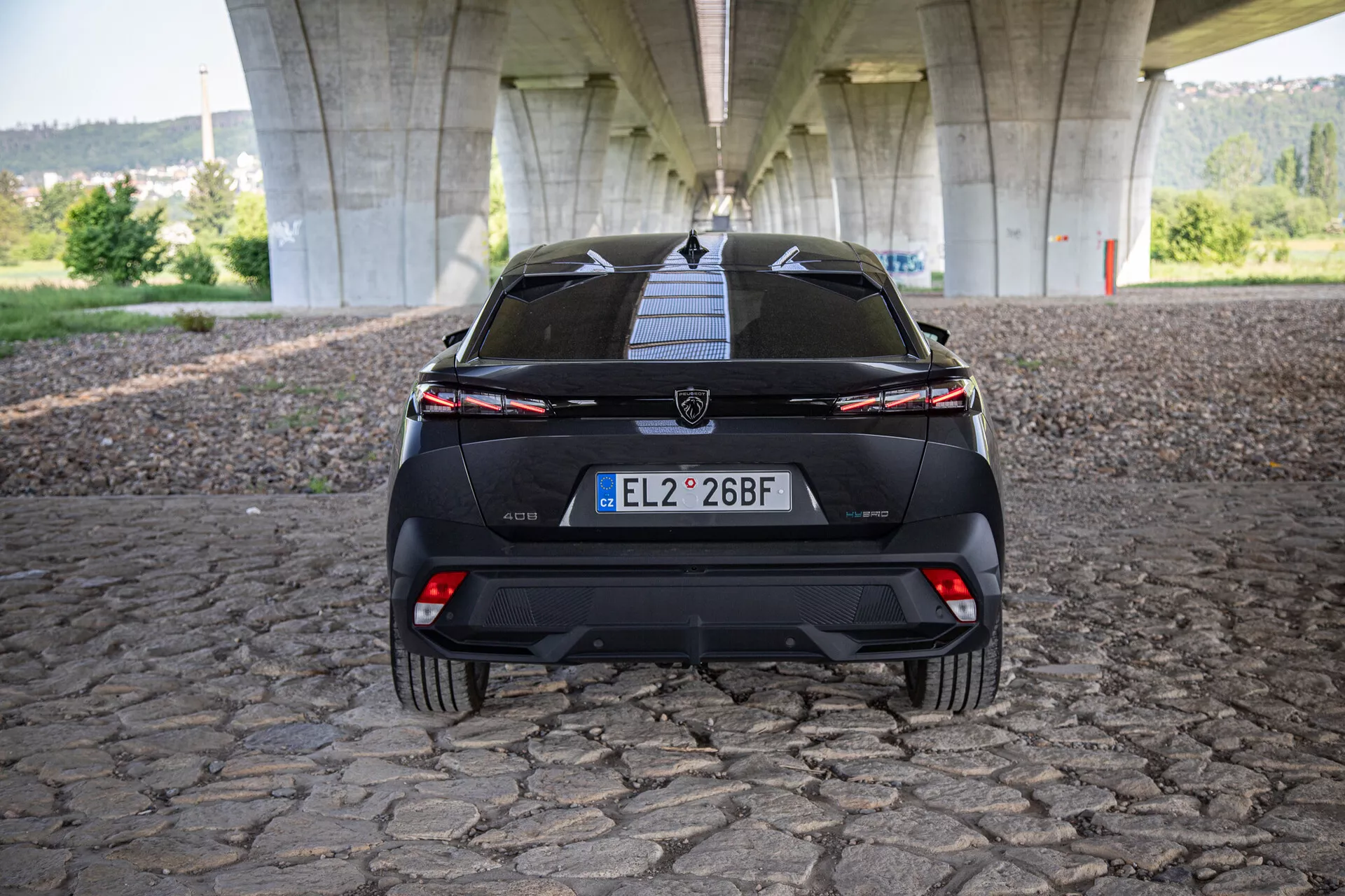 Tylny widok Peugeot 408 2023 pokazujący klapę bagażnika, zderzak, dolny difuzor, tylne światła z łączącą się belką świetlną oraz czujniki pomocy przy parkowaniu, sfotografowany pod mostem na brukowanej powierzchni