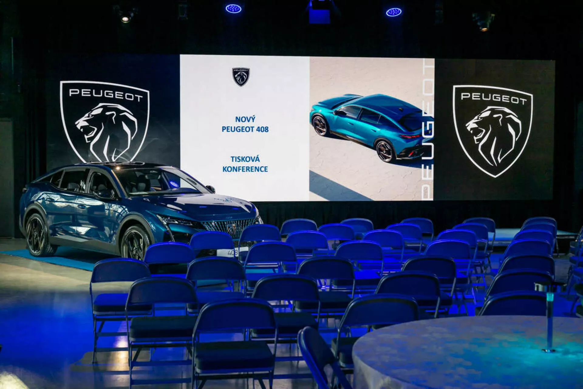 Peugeot 408 prezentowany na konferencji prasowej, widziany pod kątem 45 stopni podkreślający jego przedni i boczny profil, elegancki design oraz reflektory LED, z cyfrowymi ekranami i brandingiem w tle