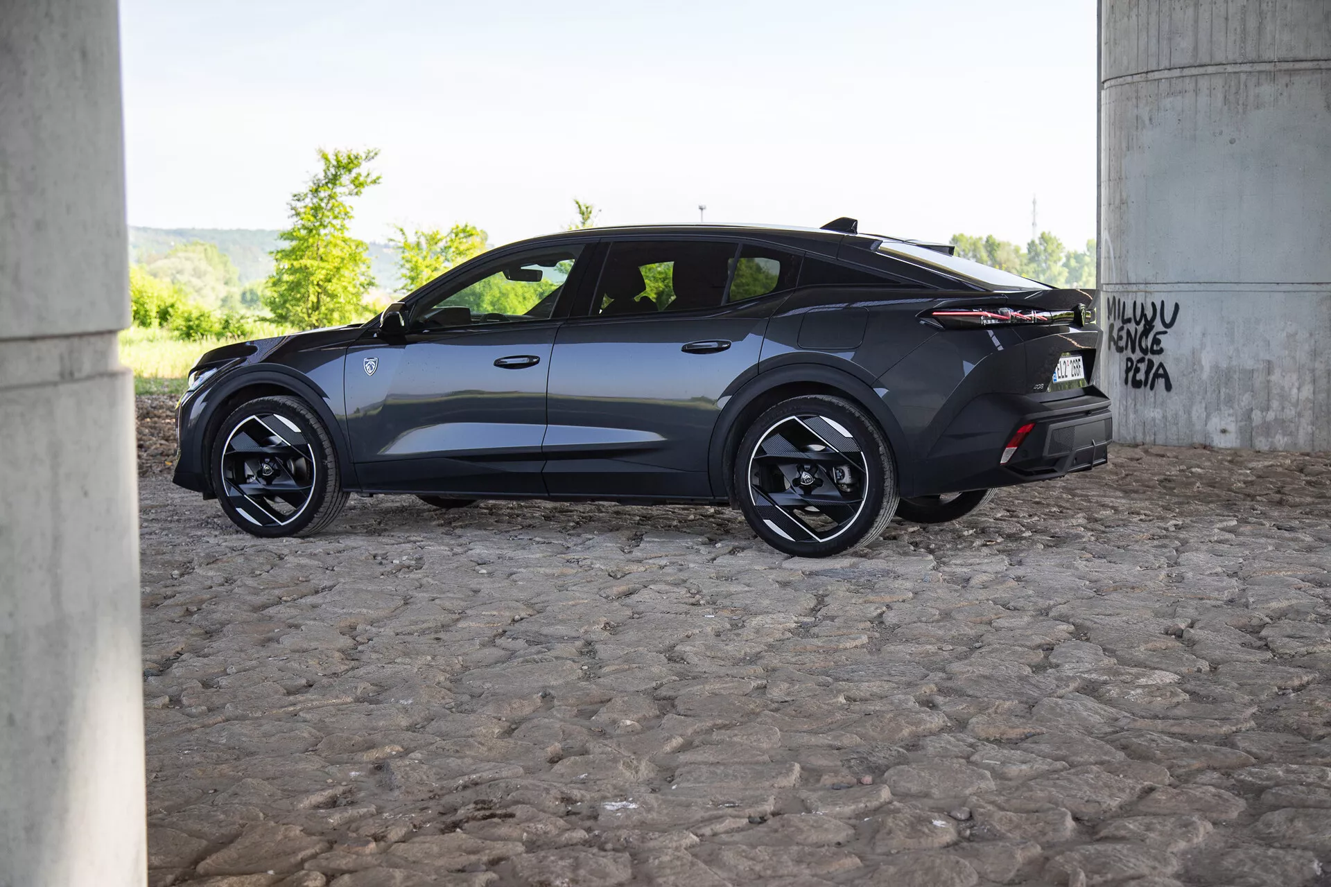 Widok z profilu nowoczesnego ciemnego samochodu Peugeot 408 2023 pod mostem z graffiti, cechujący się aerodynamicznym designem, kołami ze stopów i przyciemnianymi szybami