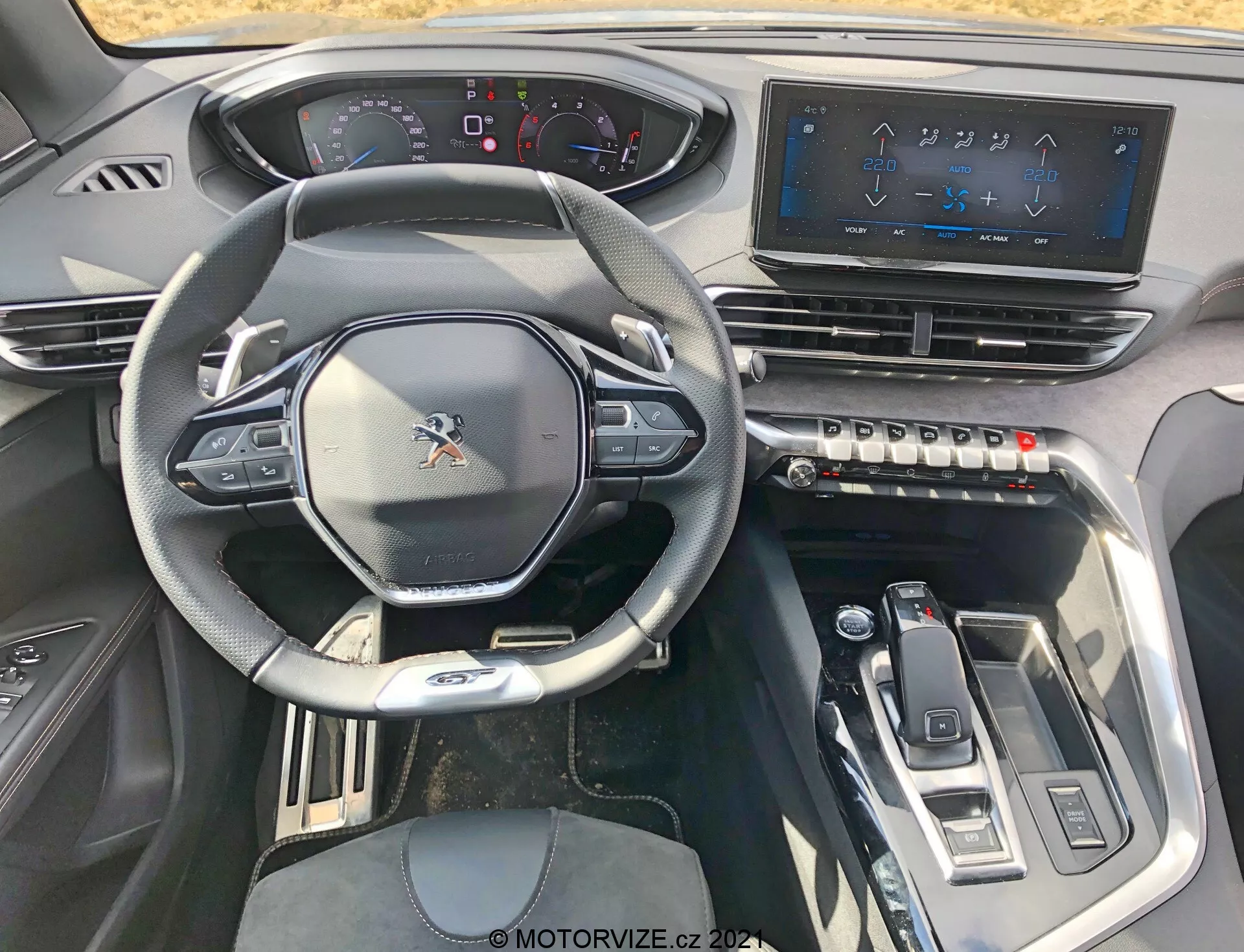 Widok wnętrza Peugeot 5008 z 2021 roku z perspektywy siedzenia kierowcy, pokazujący sportową wielofunkcyjną kierownicę z zamontowanymi przyciskami, częściowo cyfrowy zestaw wskaźników, duży dotykowy system infotainment, dotykowe przyciski klimatyzacji, elegancki selektor biegów i nowoczesne elementy wzornicze z metalicznymi akcentami, zapewniające zaawansowane technologicznie, ekskluzywne wrażenia.
