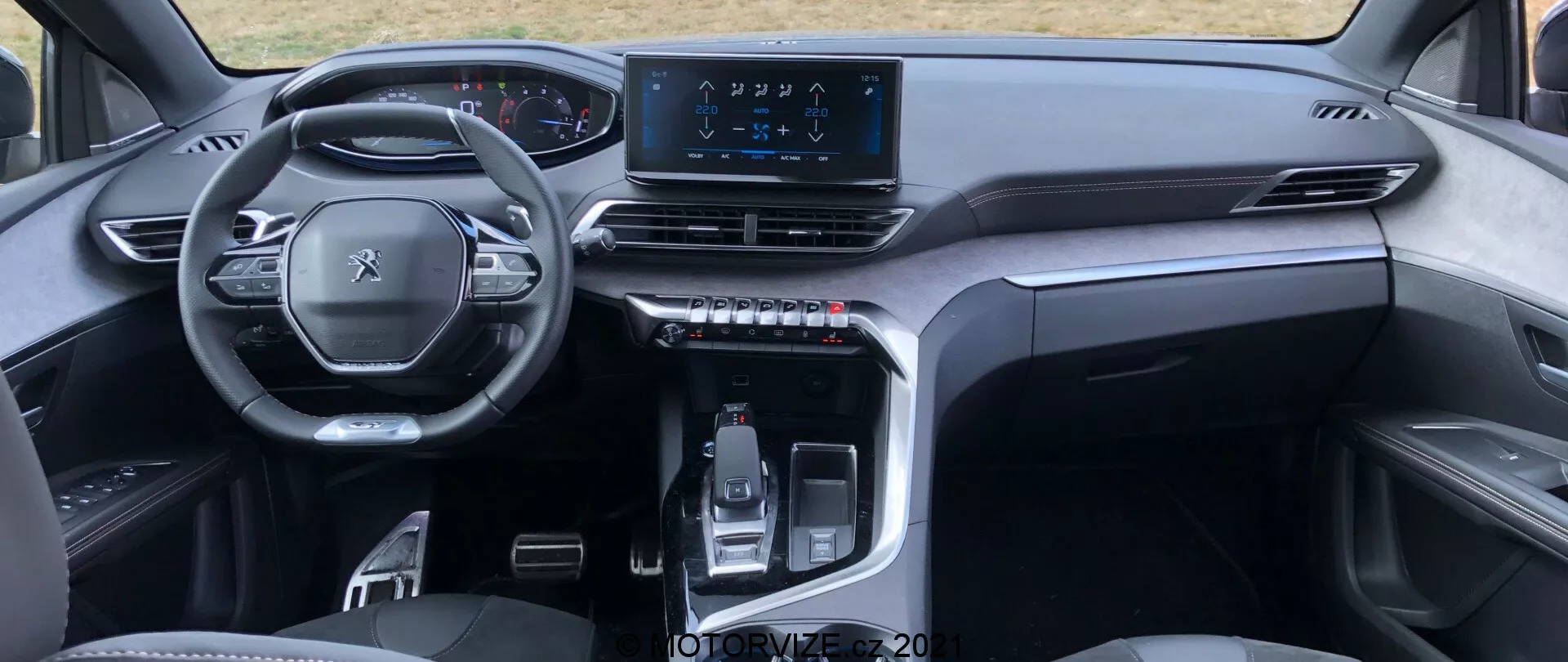 Widok wnętrza Peugeot 5008 2021 z perspektywy tylnego siedzenia, podkreślający wielofunkcyjną kierownicę, cyfrowy zestaw wskaźników, symetryczną deskę rozdzielczą z nawiewami powietrza, centralny ekran dotykowy infotainment oraz elegancką konsolę środkową z elektronicznie sterowaną dźwignią zmiany biegów i przyciskami dotykowymi. Stylowy, minimalistyczny design wykorzystuje materiały wysokiej jakości, sugerując nowoczesny, premium pojazd.