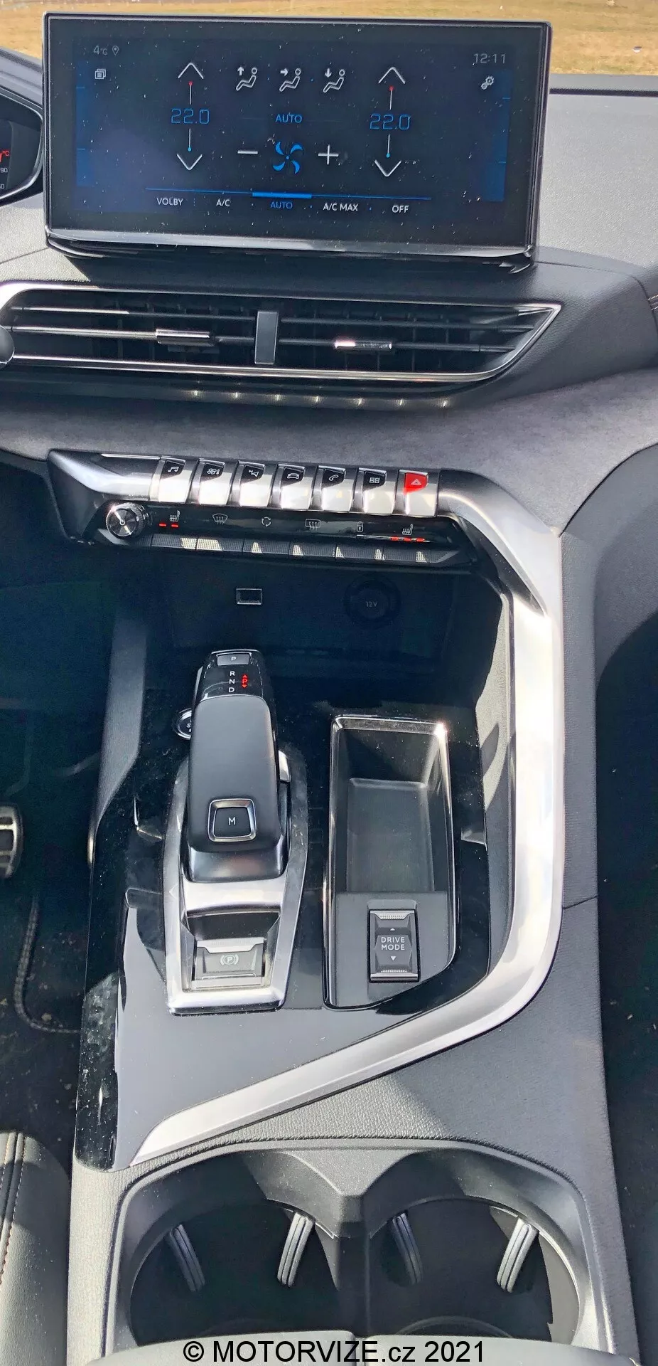 Widok wnętrza Peugeot 5008 z 2021 roku ukazujący konsolę centralną z perspektywy z góry. Obraz przedstawia unoszący się dotykowy ekran systemu multimedialnego, pokazujący sterowanie klimatyzacją nad fizycznymi przyciskami i pokrętłami do regulacji klimatu. Niżej widoczna jest elegancka elektroniczna dźwignia zmiany biegów oraz otaczające ją kontrolki do wyboru trybów jazdy i wielofunkcyjne pokrętło interfejsu. Design sugeruje nowoczesny, najwyższej klasy pojazd z zaawansowanymi technologiami oraz ergonomicznymi funkcjami, w tym uchwytami na napoje i przestrzeniami do przechowywania.